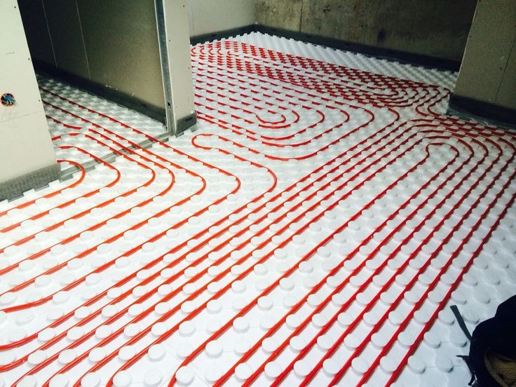 Montaż czerwonych rur na białych płytach izolacyjnych, tworzących system rozprowadzania ciepła pod podłogą w surowym pomieszczeniu.