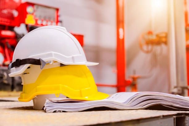 Dwa kaski budowlane, biały i żółty, leżące na stercie dokumentów na tle czerwonej konstrukcji i rozproszonego światła.