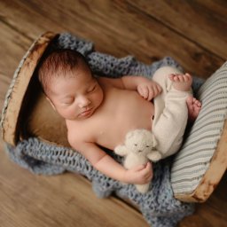 Zaczarowana Migawka studio fotograficzne - Śpiący noworodek w miniaturowym drewnianym łóżeczku na tle drewnianej podłogi, trzymający małego pluszowego misia.