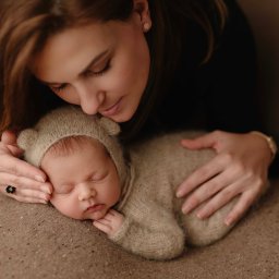 Zaczarowana Migawka studio fotograficzne - Matka czule spogląda na śpiące nowonarodzone dziecko ubrane w dzianinowy kombinezon z uszkami, leżące na teksturowanym tle; kobieta ma pierścionek z czarnym oczkiem.