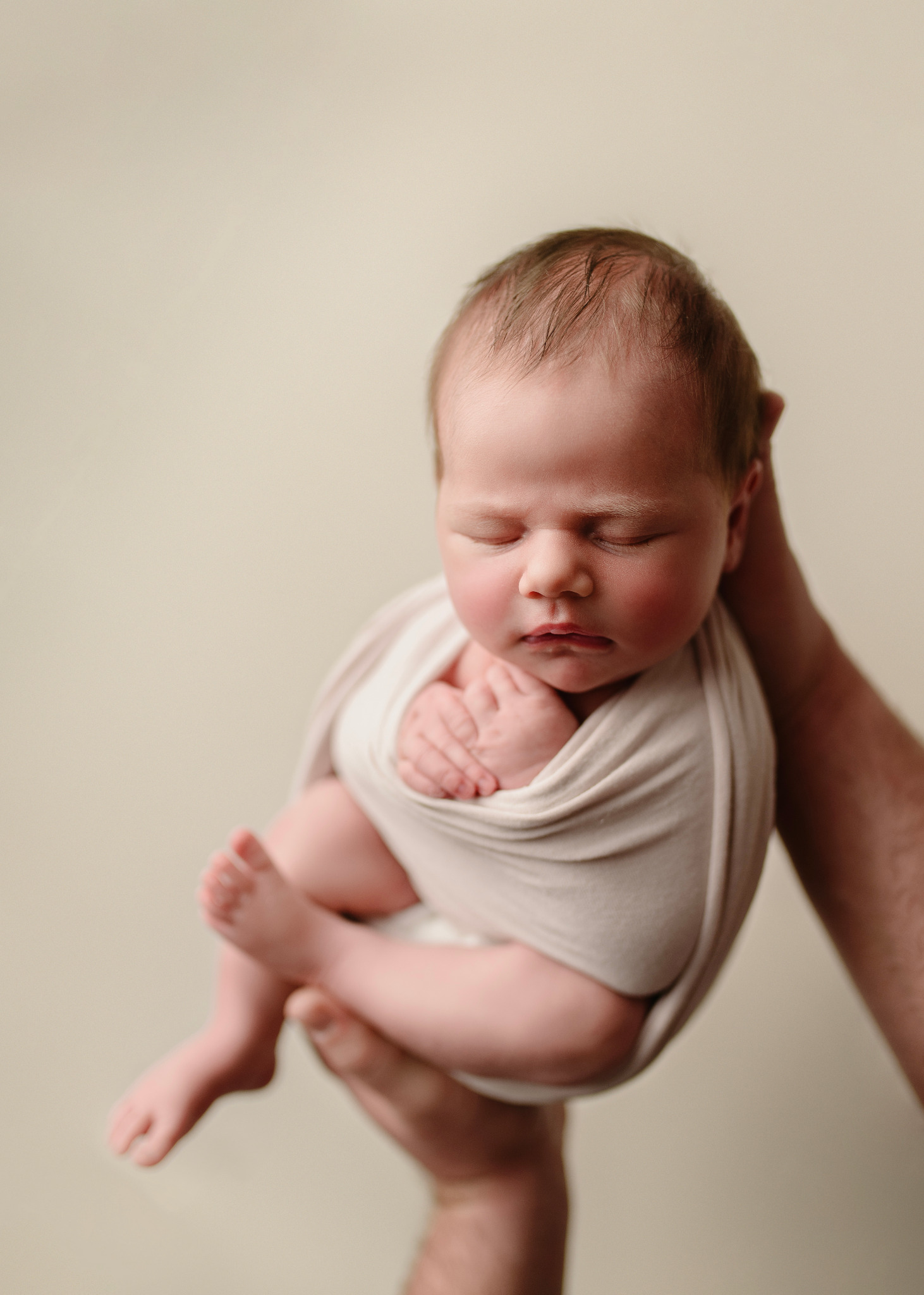 Noworodek zawinięty w beżowy materiał, trzymany w dłoniach na jasnym tle, z widocznymi drobnymi włoskami na głowie i zaciśniętymi piąstkami.