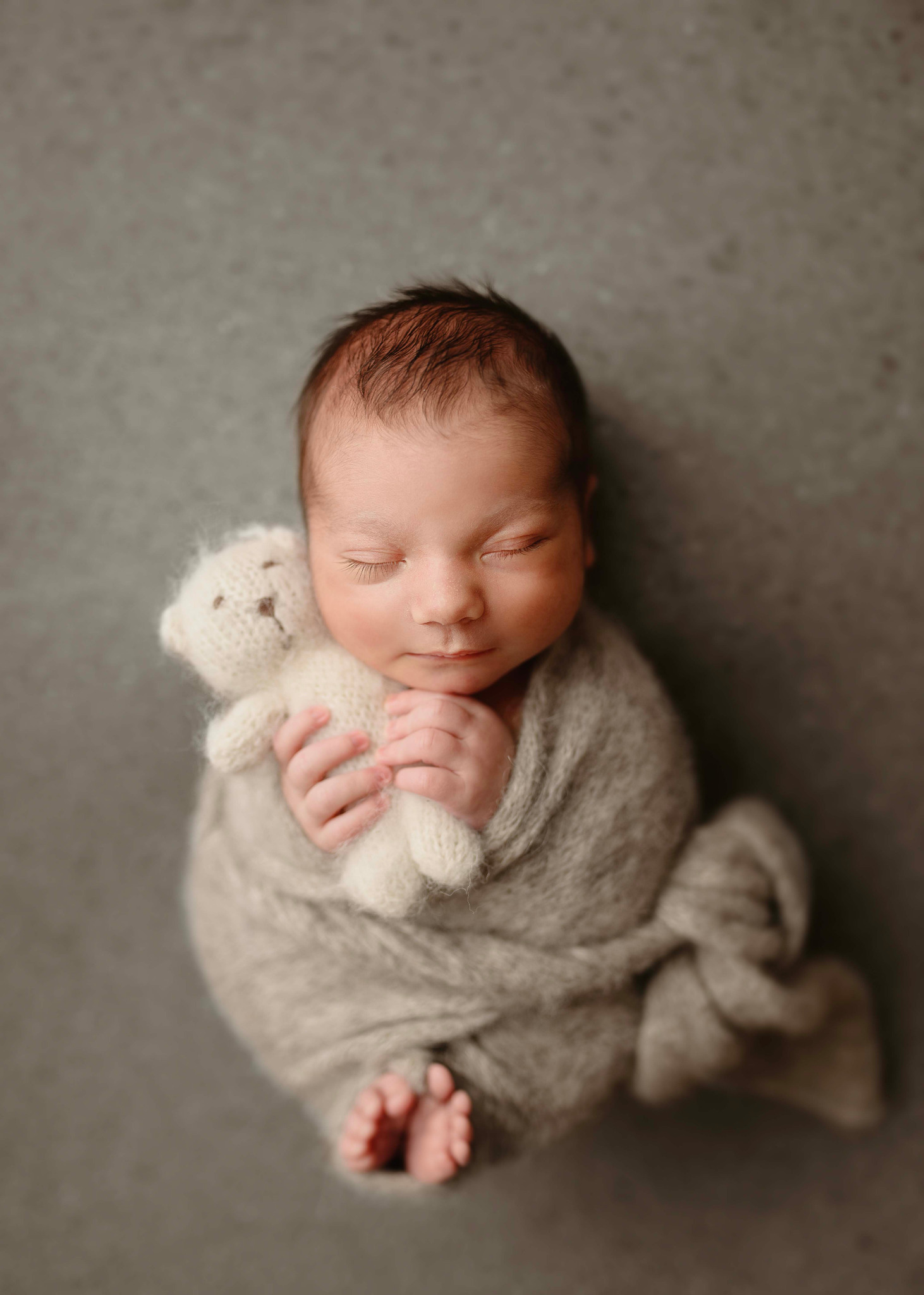 Noworodek owinięty w szary pled śpiący na szarym tle, trzymający w rączkach małego pluszowego misia.