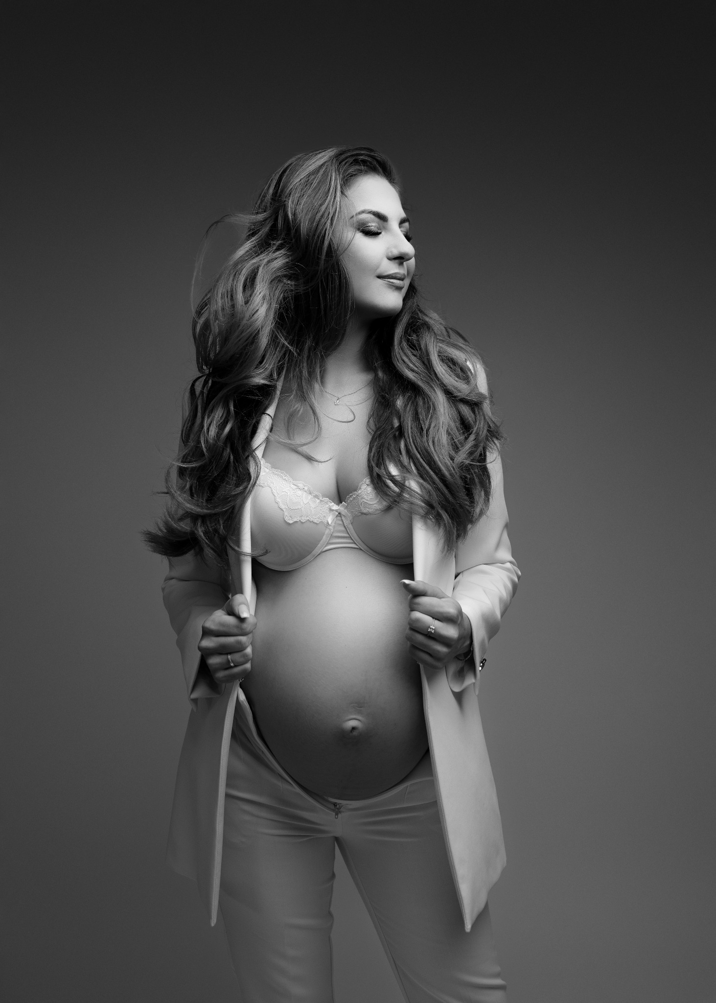 Czarno-białe portretowe ujęcie kobiety w ciąży, z długimi falowanymi włosami, w rozpiętym kremowym żakiecie i biustonoszu z koronką, na jednolitym szarym tle.