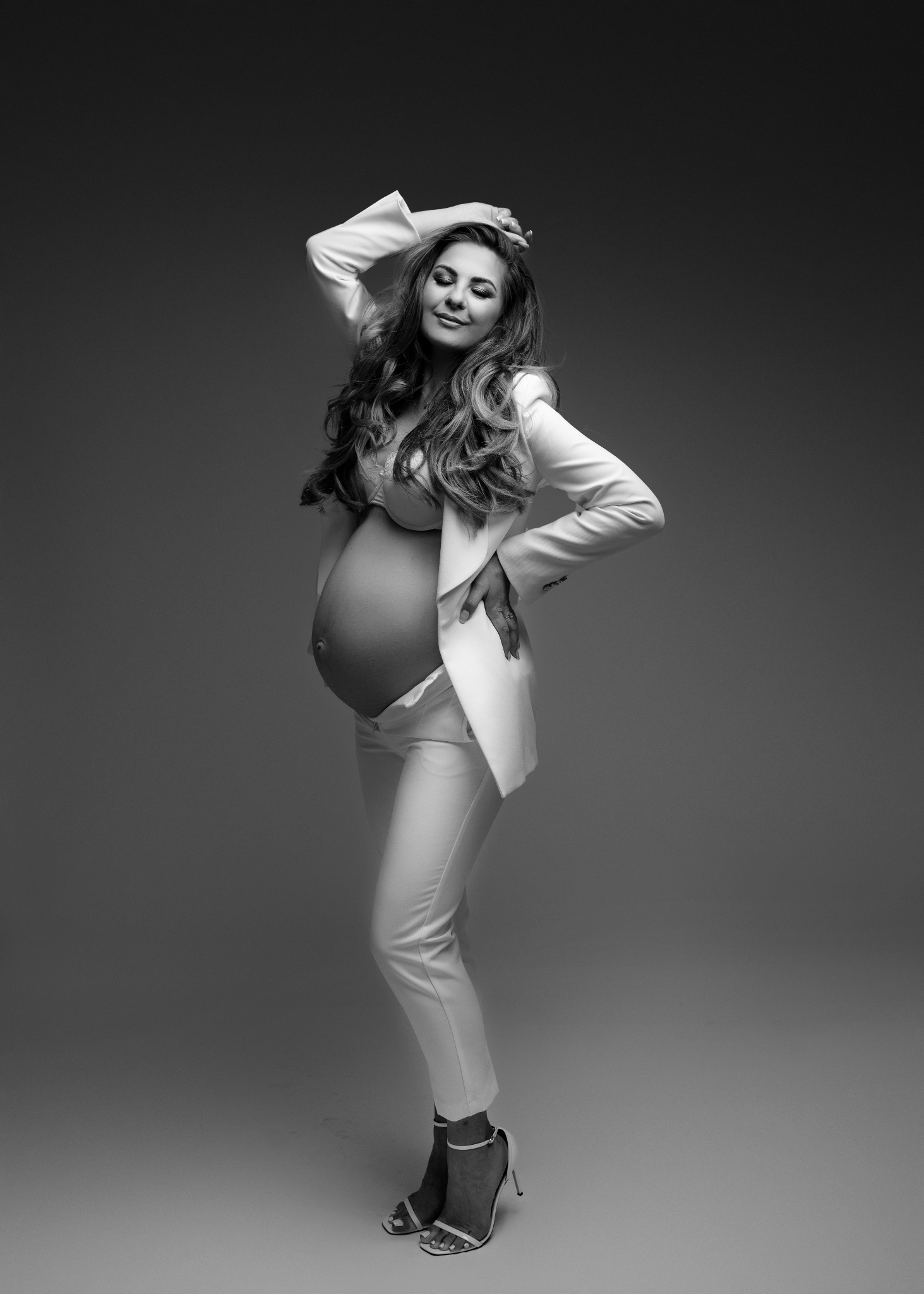 Czarno-białe zdjęcie kobiety w ciąży, pozującej w białym garniturze i szpilkach na szarym tle, z ręką uniesioną nad głową i zamkniętymi oczami.