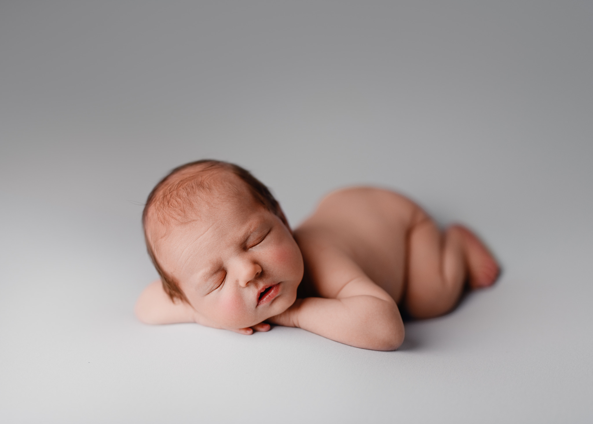 Śpiący noworodek ułożony na brzuszku z rączkami pod policzkiem, na gładkim, jasnoszarym tle.
