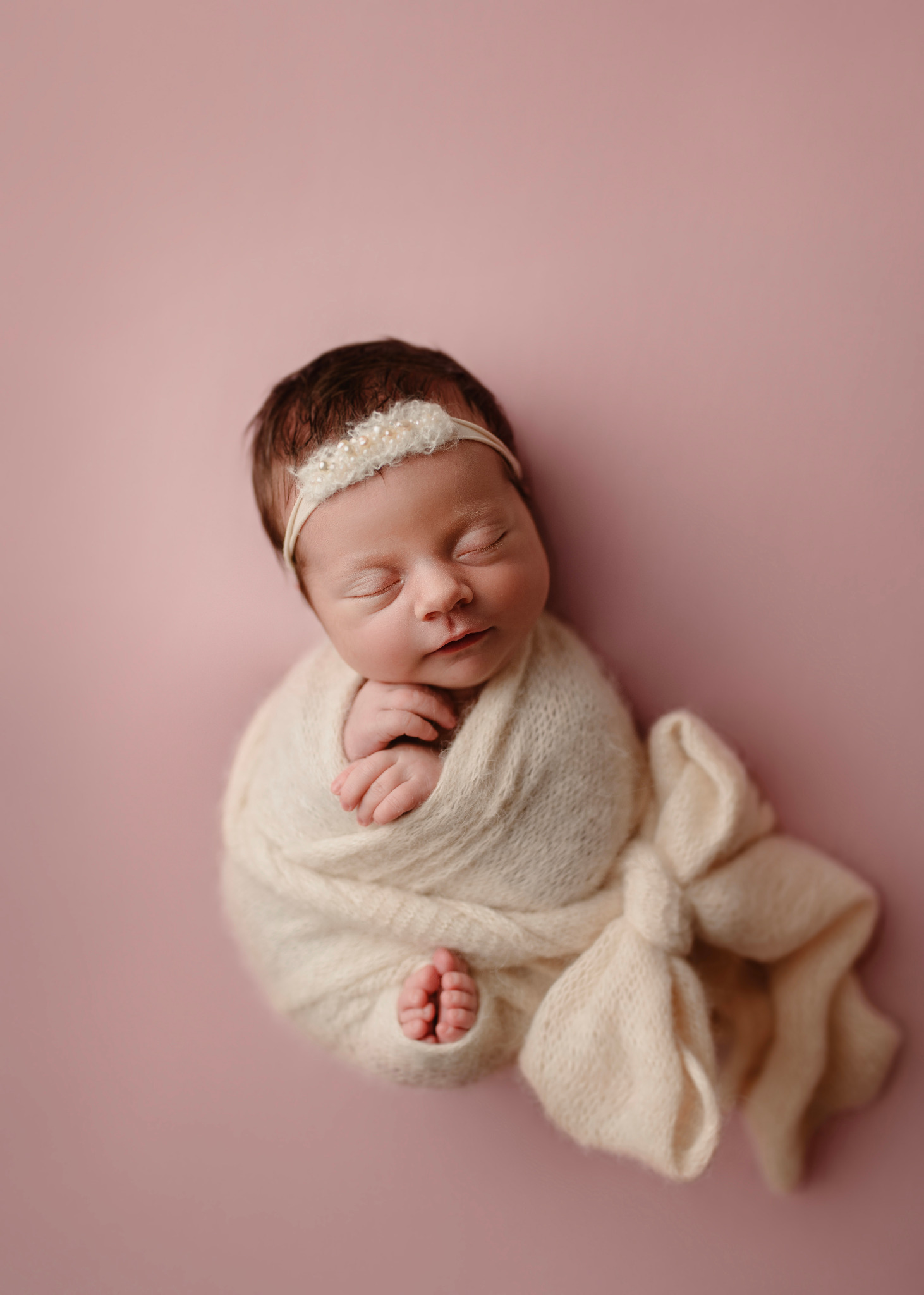 Śpiący noworodek zawinięty w kremowy, dziergany kocyk z kokardą, leżący na różowym tle, z opaską z perełkami na głowie.