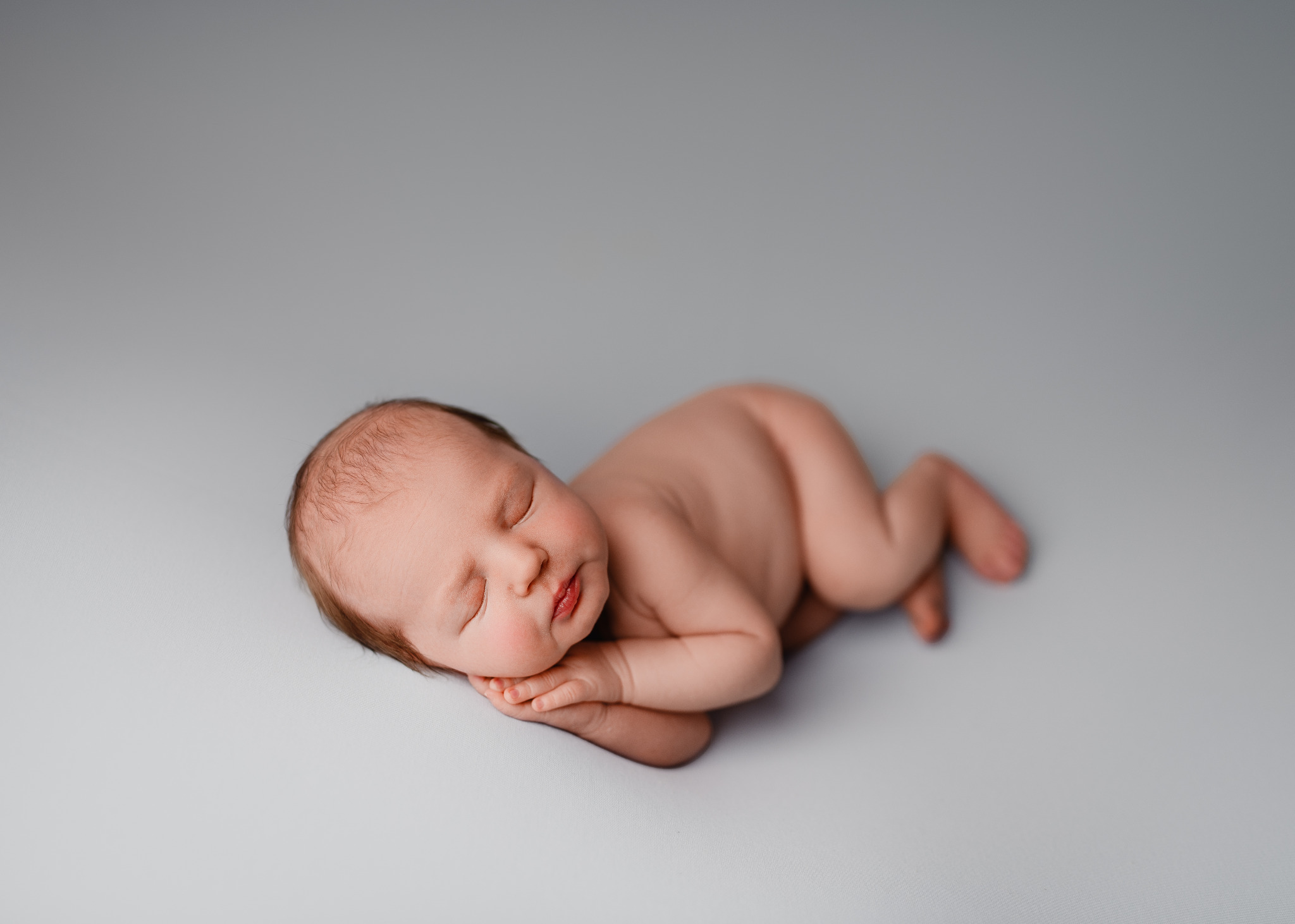 Noworodek śpiący na boku, z delikatnie zaczerwienionymi usteczkami, ułożony na gładkim, jasnoszarym tle.