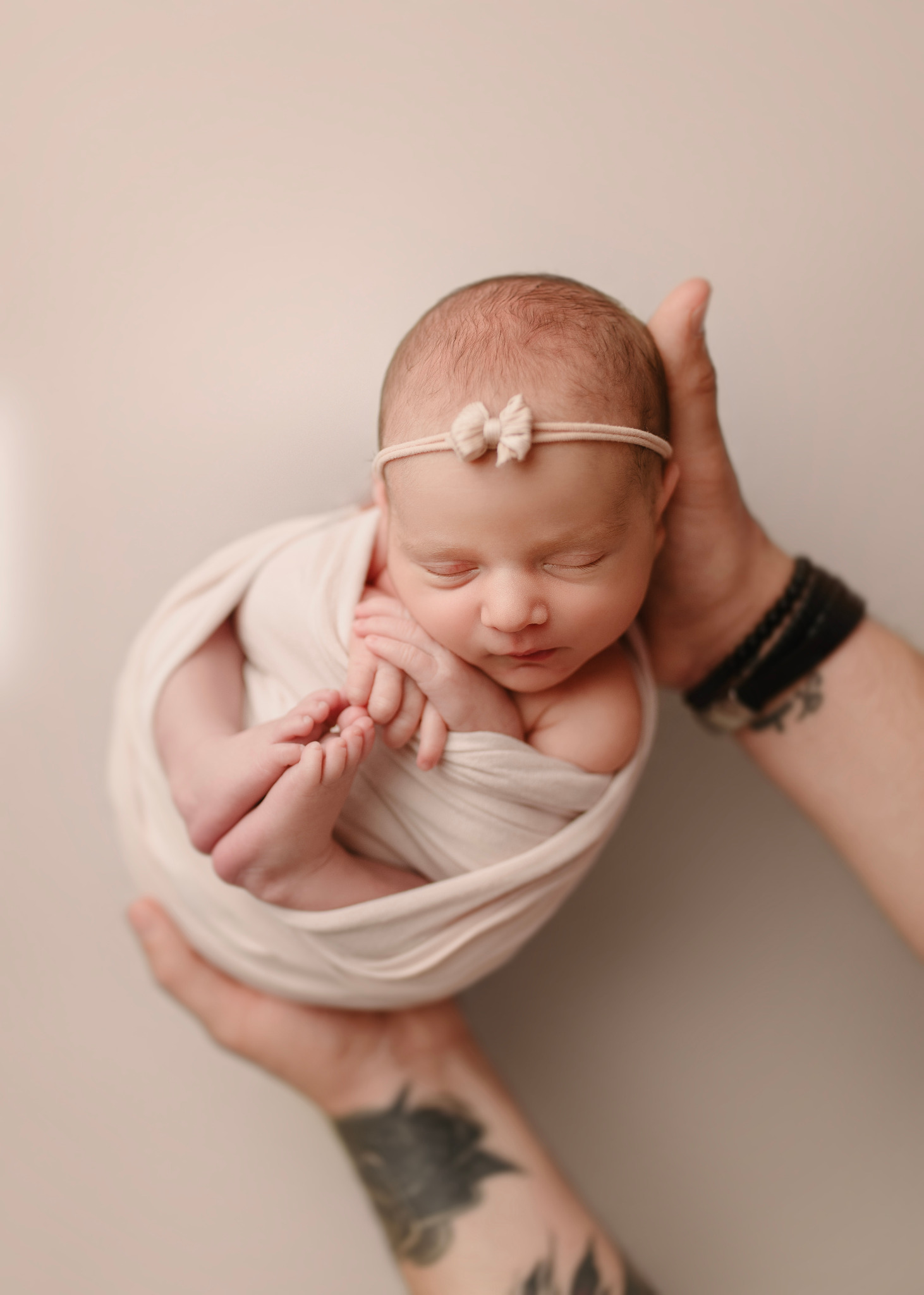 Śpiące niemowlę zawinięte w kremowy materiał, z małą kokardką na głowie, delikatnie trzymane w dłoniach osoby z tatuażem na przedramieniu i skórzanymi bransoletkami.