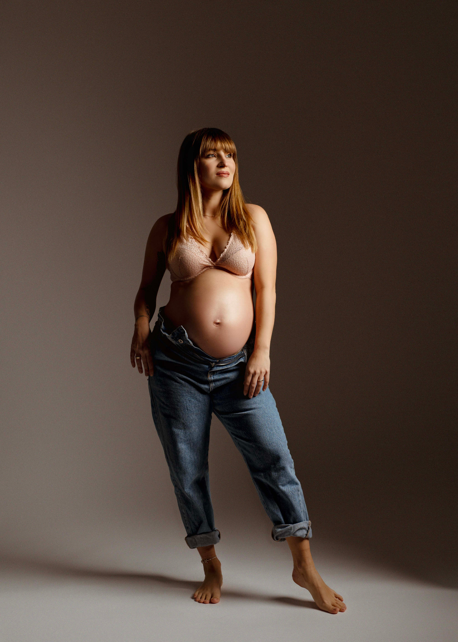 Kobieta w ciąży, w dżinsach i koronkowym biustonoszu, pozuje na jednolitym tle, spoglądając w lewo, z widocznym tatuażem na przedramieniu i pierścionkiem na palcu.