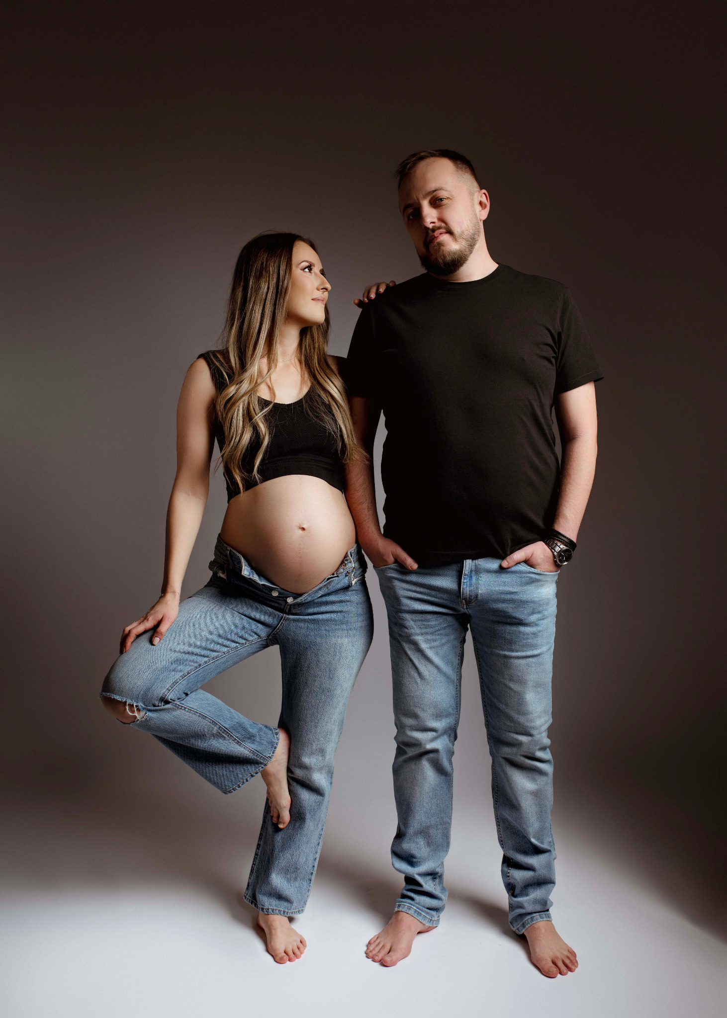 Kobieta w rozpiętych jeansach i czarnym topie, eksponująca ciążowy brzuszek, opiera stopę o kolano, spoglądając na mężczyznę w czarnej koszulce i jeansach, stojącego obok z rękami w kieszeniach...