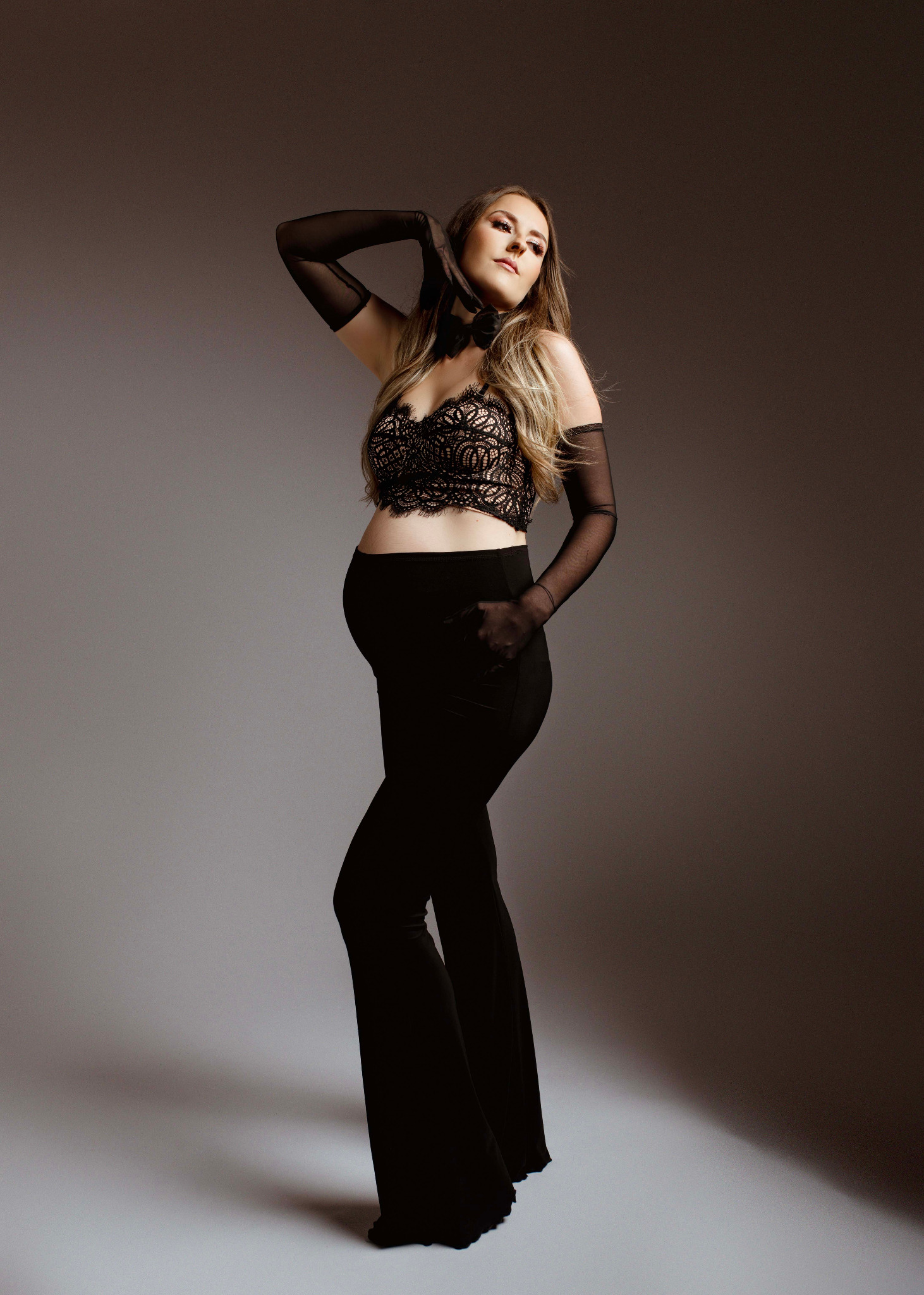 Elegancka sesja ciążowa: Kobieta w czarnej koronkowej bieliźnie i długich rękawiczkach pozuje na szarym tle, dłonie uniesione do góry.