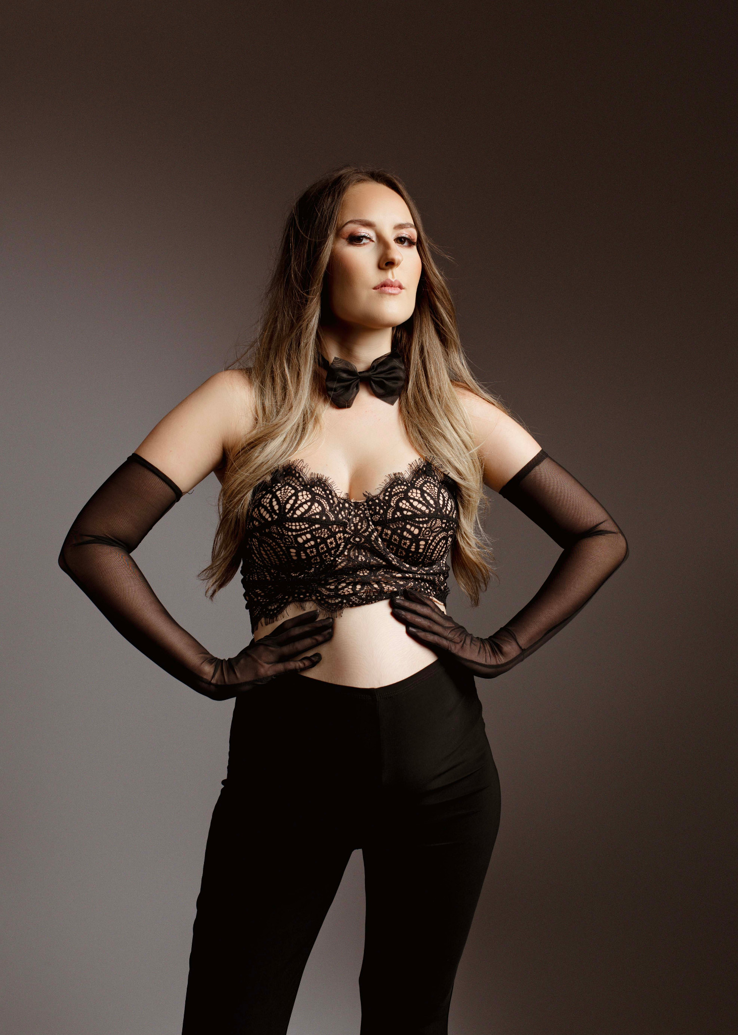 Kobieta w czarnym koronkowym topie, muszce i długich rękawiczkach pozuje z rękami na biodrach na szarym tle.