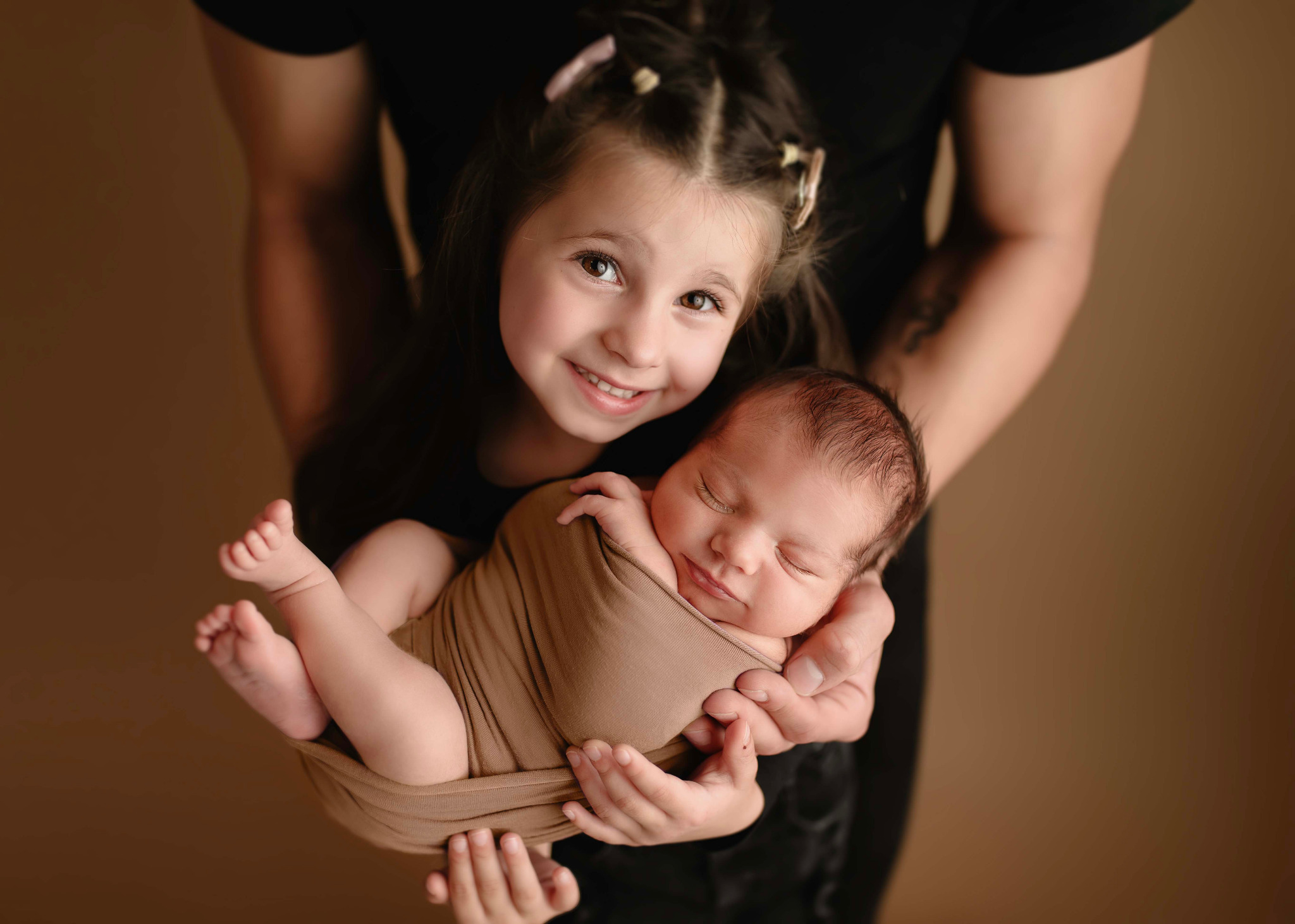Uśmiechnięta dziewczynka trzyma w ramionach śpiącego noworodka zawiniętego w brązowy materiał, na beżowym tle, widoczne dłonie trzymające dziecko.