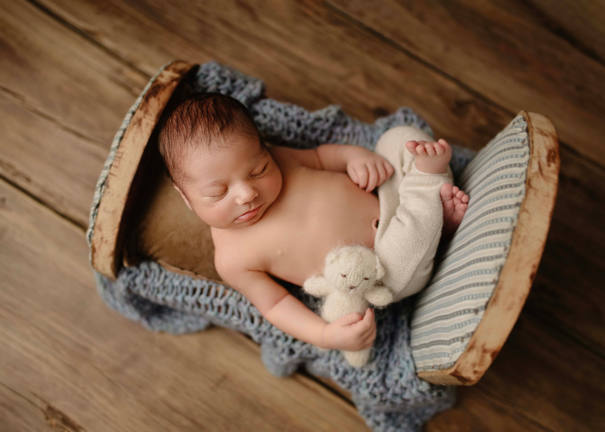 Śpiący noworodek w miniaturowym drewnianym łóżeczku na tle drewnianej podłogi, trzymający małego pluszowego misia.