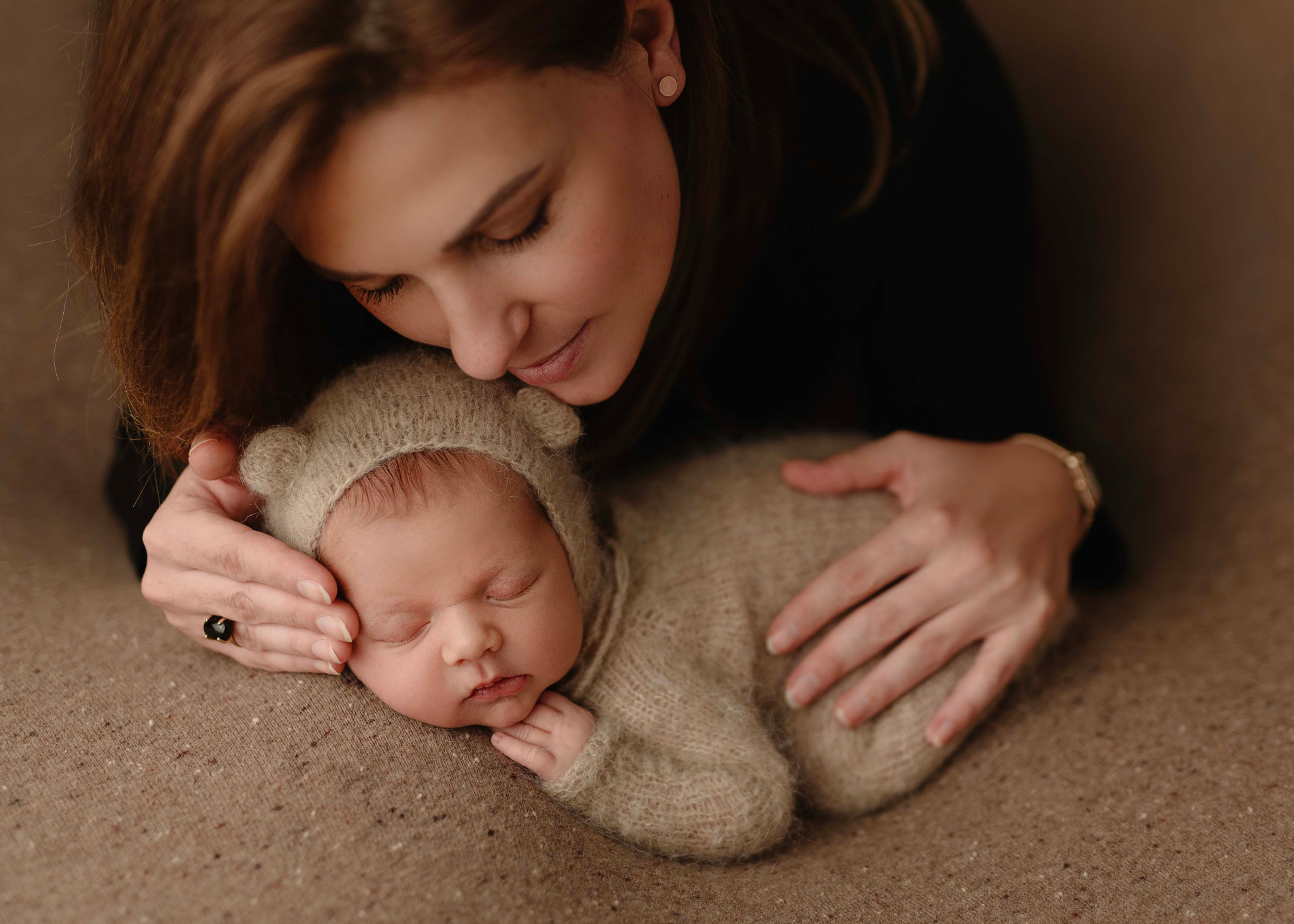 Matka czule spogląda na śpiące nowonarodzone dziecko ubrane w dzianinowy kombinezon z uszkami, leżące na teksturowanym tle; kobieta ma pierścionek z czarnym oczkiem.
