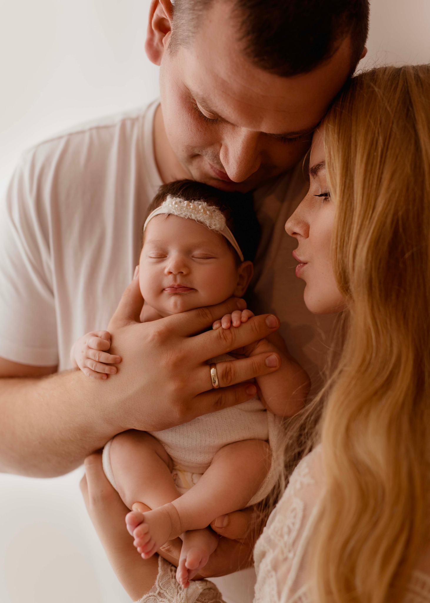 Rodzinne ujęcie: śpiąca noworodka z opaską na głowie, trzymana przez rodziców, z widocznym złotym pierścionkiem na palcu ojca.