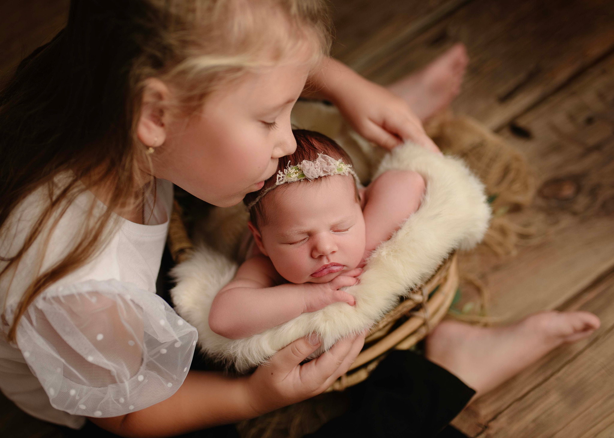 Dziewczynka całuje śpiącego noworodka z opaską w wianuszku, ułożonego w koszyku wyłożonym białym futerkiem na drewnianej podłodze.