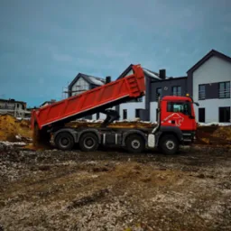 Czerwona wywrotka MAN z podniesioną skrzynią ładunkową wysypuje ziemię na placu budowy, w tle domy w zabudowie szeregowej i pochmurne niebo.