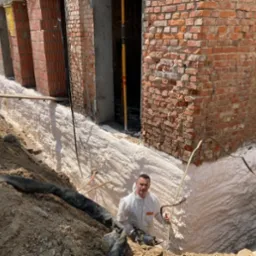 Pracownik w kombinezonie ochronnym natryskuje białą pianę izolacyjną na fundament budynku z czerwonej cegły, widoczna warstwa izolacji na ścianach wykopu.