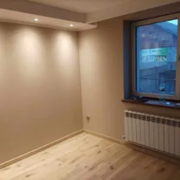 Pomalowane na beżowo wnętrze pokoju z panelami podłogowymi, białym grzejnikiem pod oknem i sufitowym oświetleniem punktowym.