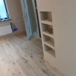 Jasne panele podłogowe z wyraźnym wzorem drewna, w tle fragment ściany z wnękami p&oacute;łkowymi i otwarte drzwi.