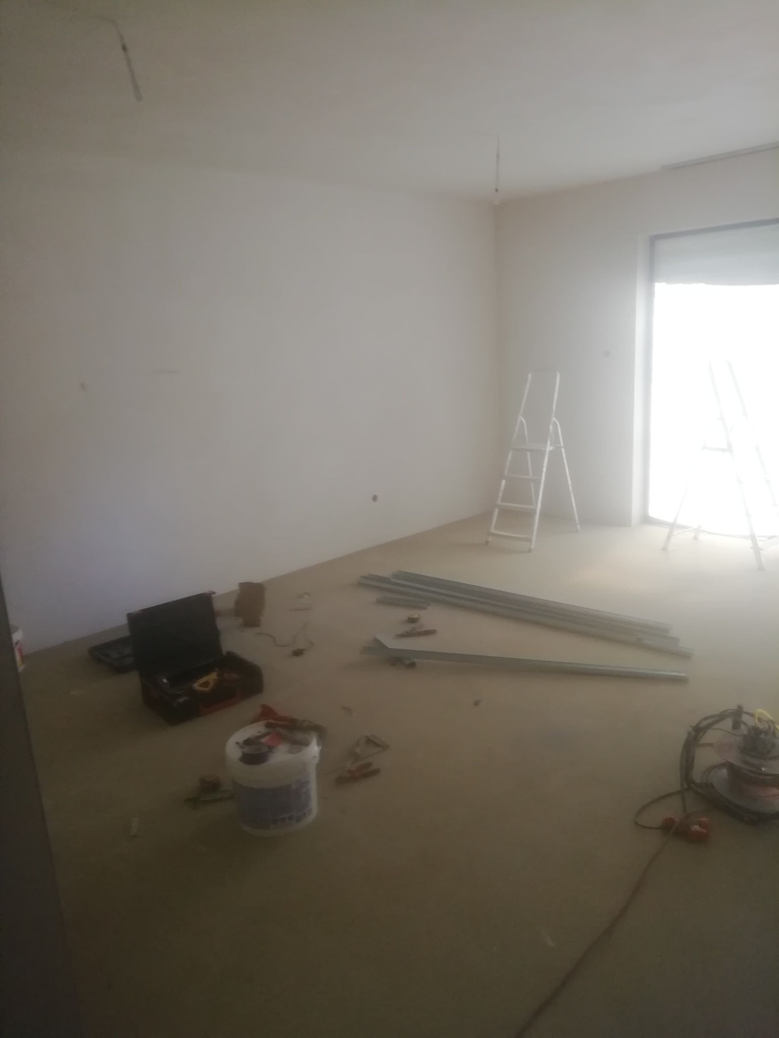 Wnętrze pokoju w trakcie remontu: widoczne profile metalowe na podłodze, drabina, narzędzia w skrzynce, wiadro z farbą i szlifierka, przygotowanie do prac wykończeniowych.