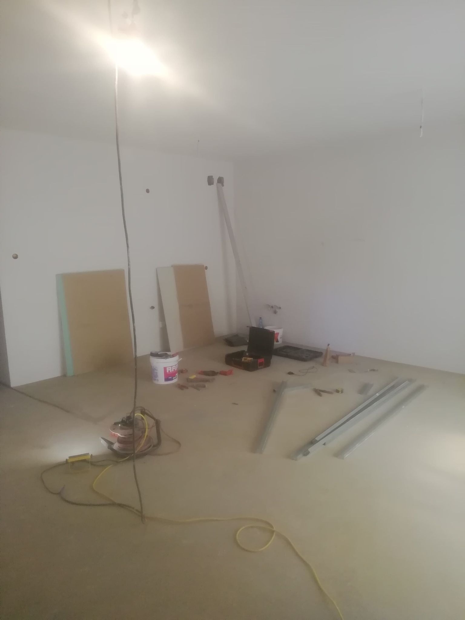Pokój w trakcie remontu: białe ściany, surowy beton na podłodze, narzędzia, puszka farby RAPID i profile aluminiowe na podłodze, widoczne ślady po instalacjach na ścianie, tymczasowe oświetlenie.
