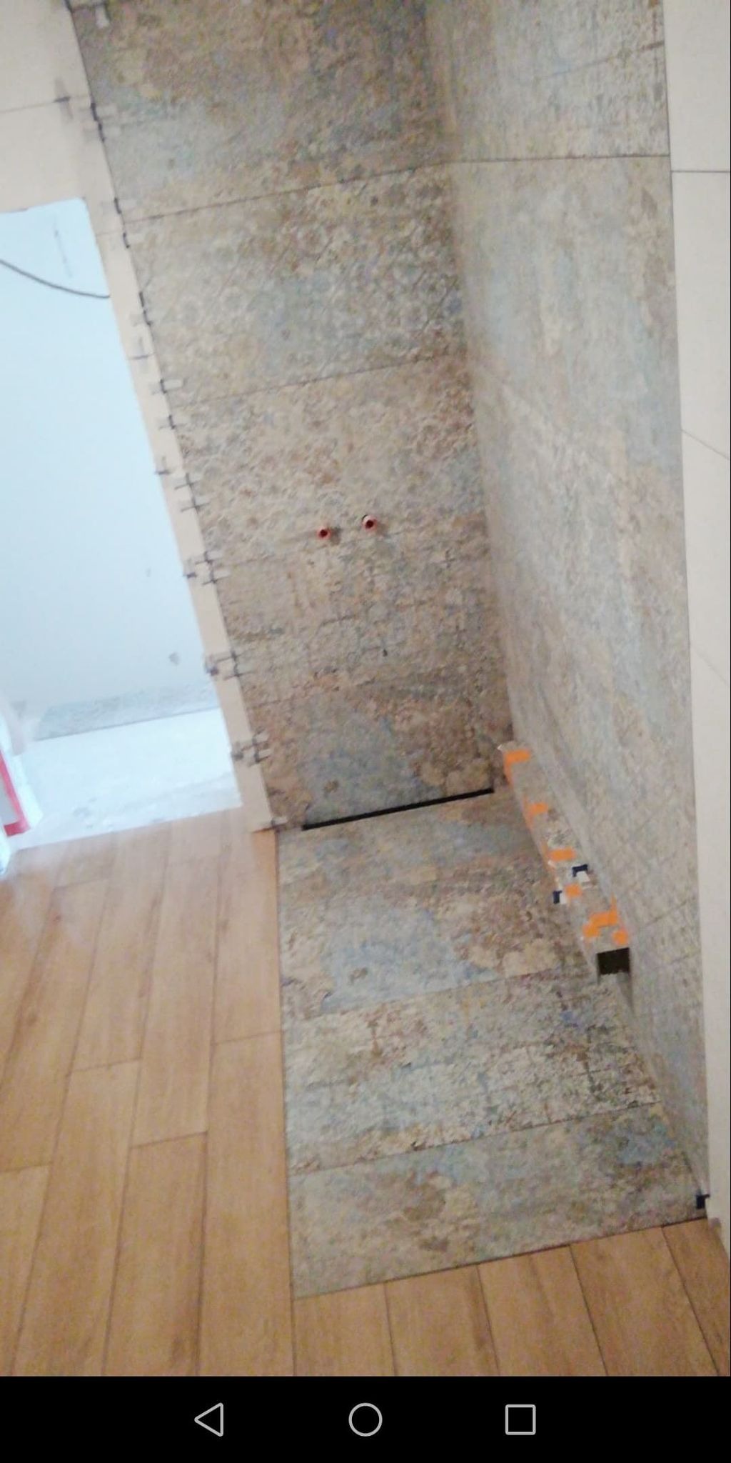 Ściana łazienki w trakcie wykańczania, z widocznymi płytkami o wzorze imitującym kamień, krzyżykami dystansowymi i fragmentem drewnopodobnej podłogi.