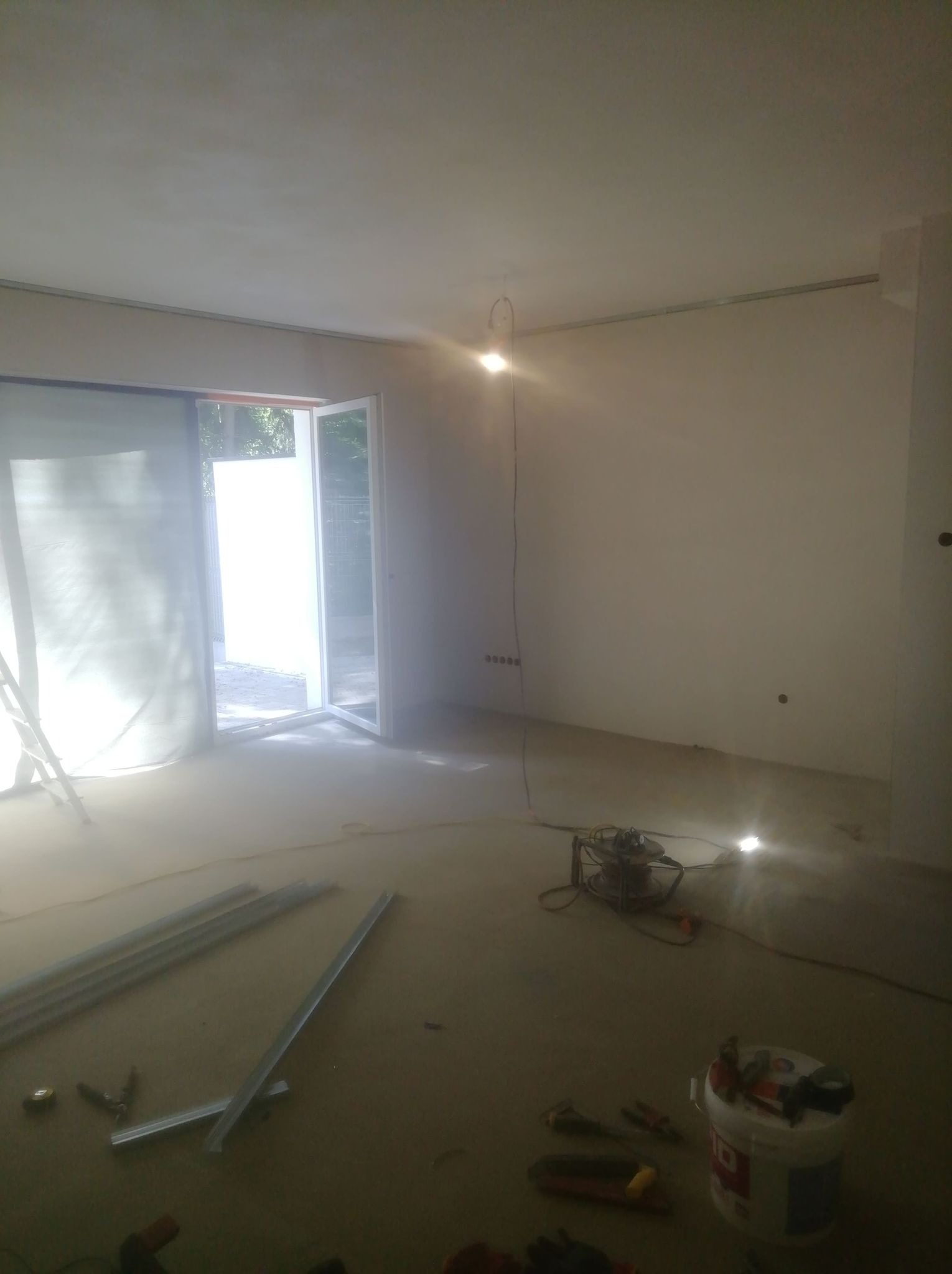 Wnętrze w trakcie remontu: surowe ściany, rozłożone profile metalowe na podłodze, narzędzia i wiadro z farbą, otwarte drzwi balkonowe z widokiem na zieleń.