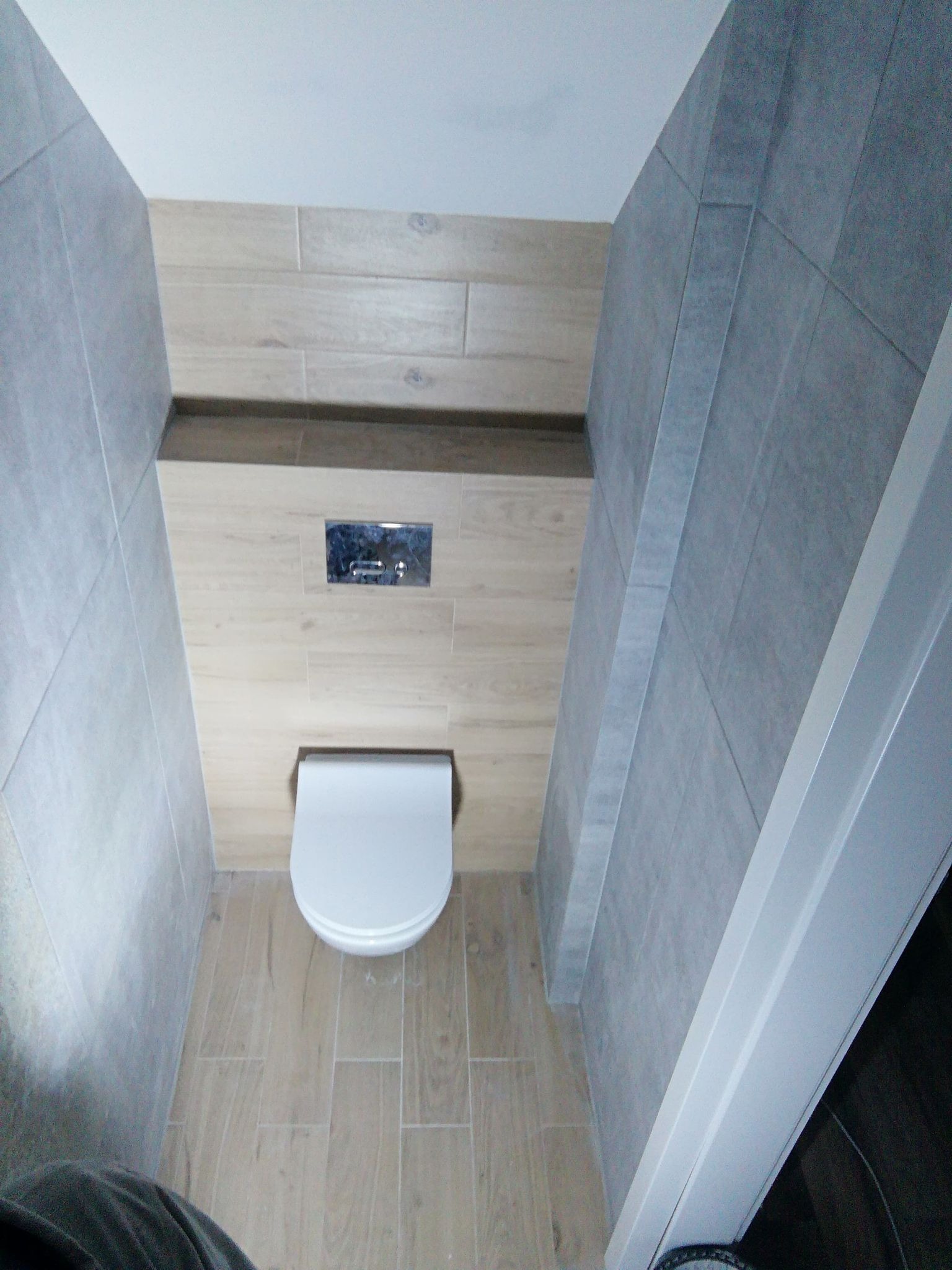 Wąska łazienka z toaletą wiszącą i spłuczką podtynkową, wykończona płytkami imitującymi drewno i szarym gresem. Widok z góry na wykończoną wnękę.