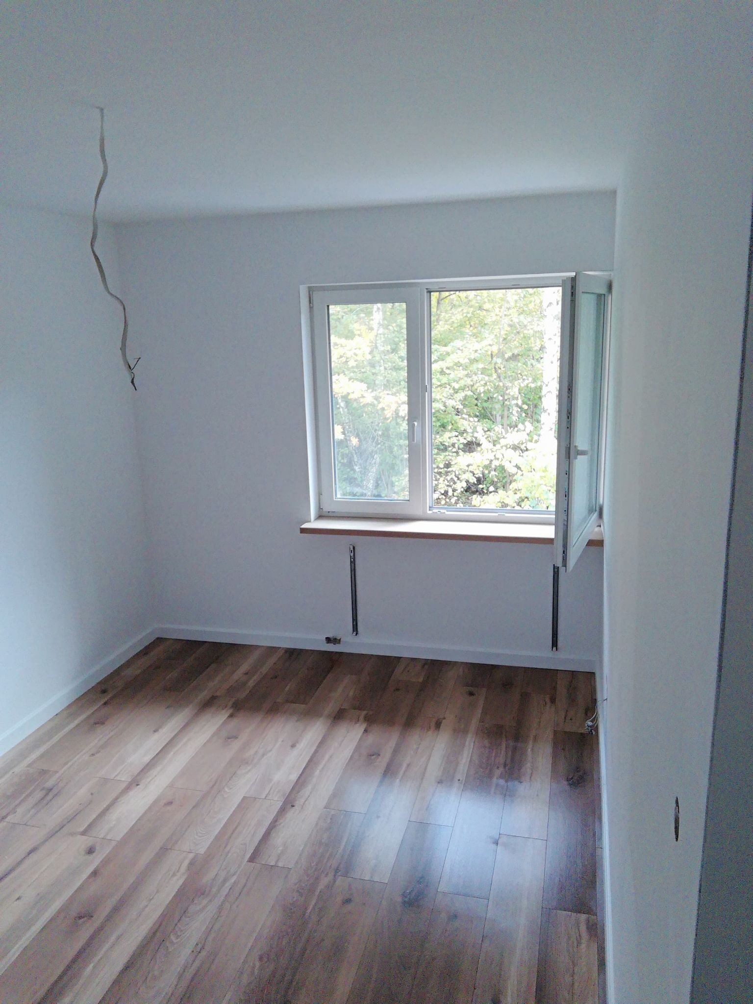 Pusty pokój po remoncie z drewnianą podłogą, białymi ścianami i otwartym oknem z widokiem na drzewa, widoczne wystające kable ze ściany i sufitu.