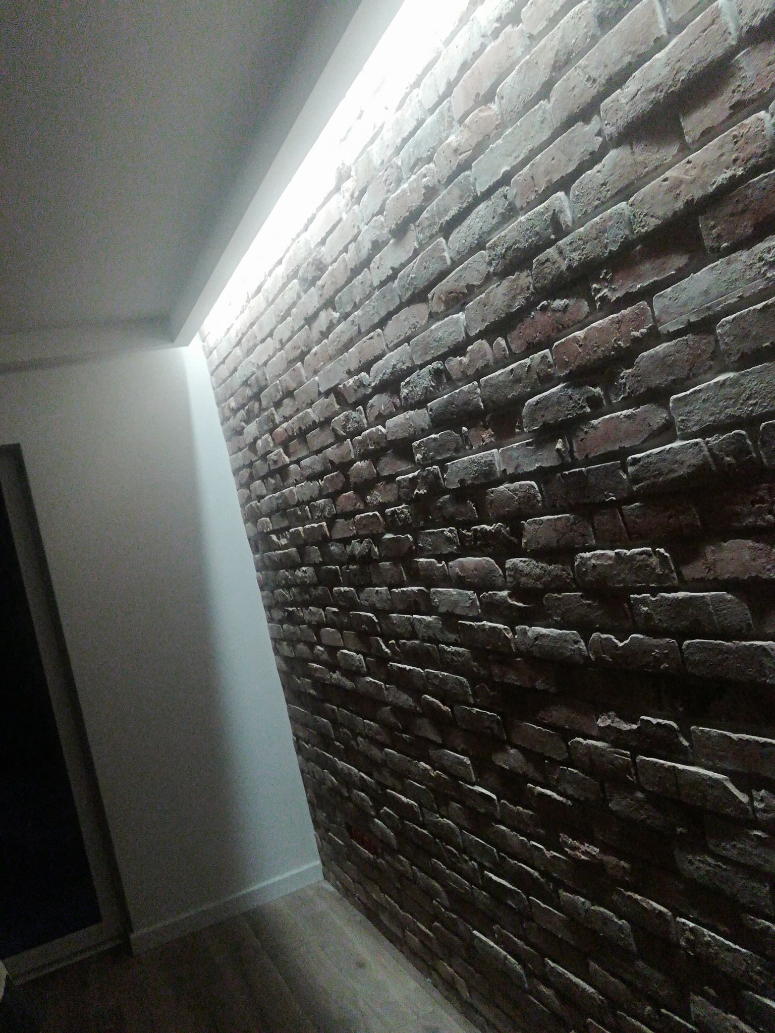 Ściana wykończona cegłą dekoracyjną z podświetleniem LED wzdłuż sufitu, widoczny fragment podłogi i drzwi.
