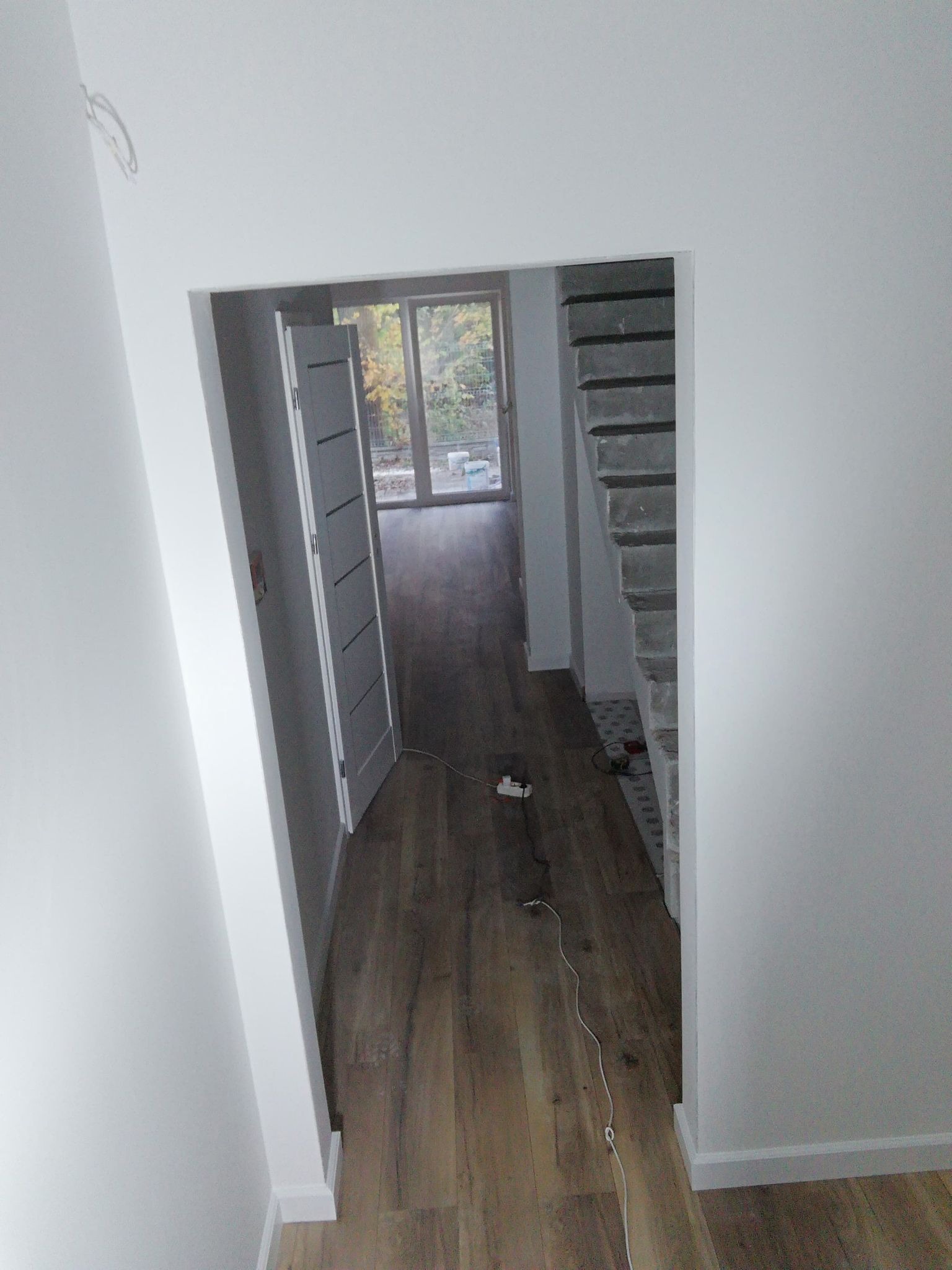 Wnętrze w trakcie remontu: świeżo pomalowane białe ściany, drewnopodobne panele podłogowe, częściowo zamontowane szare drzwi wewnętrzne, surowe betonowe schody z widoczną konstrukcją.