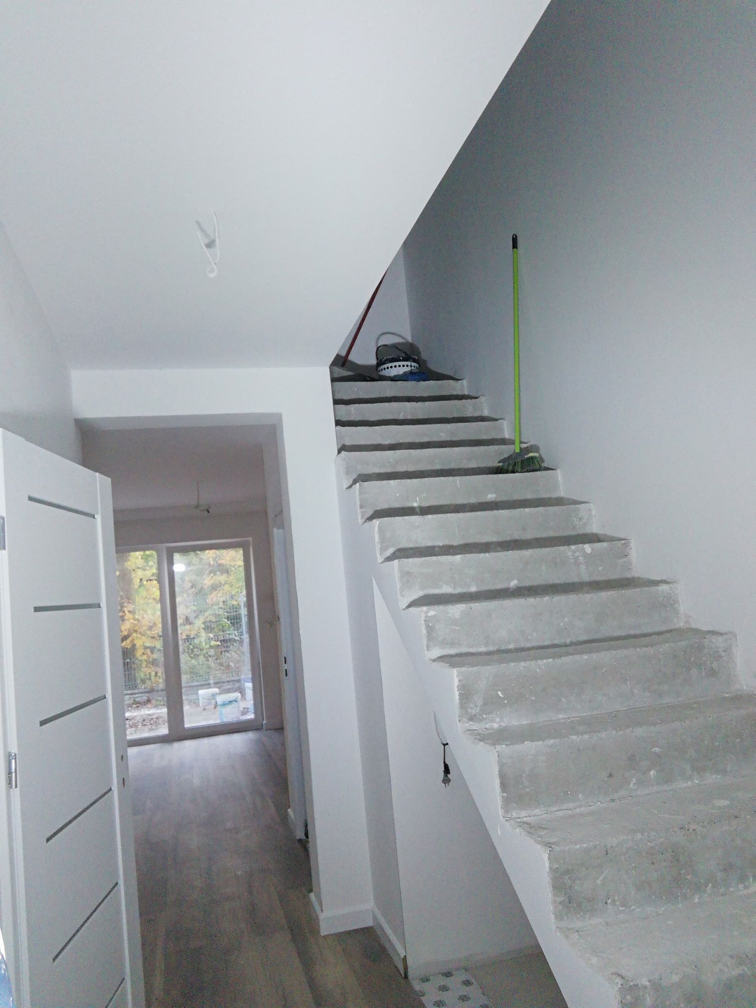 Surowe betonowe schody wewnętrzne w trakcie budowy lub remontu, z widocznymi śladami po pracach i narzędziami na stopniach, otwarte drzwi z widokiem na parkiet w tle.