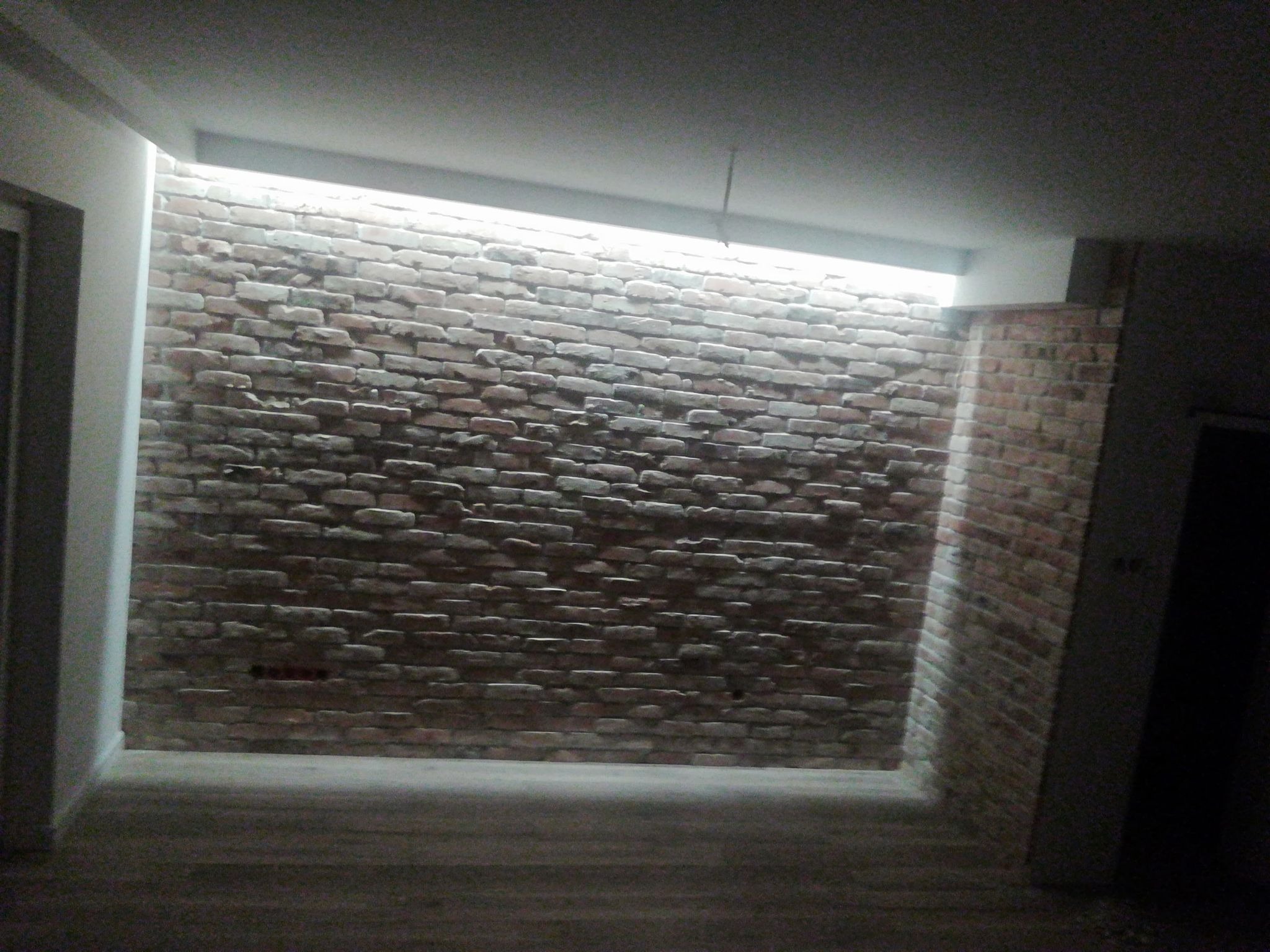Surowa ściana z cegły w korytarzu, oświetlona listwą LED w suficie, widoczny fragment sufitu i podłogi w ciemnych tonacjach.
