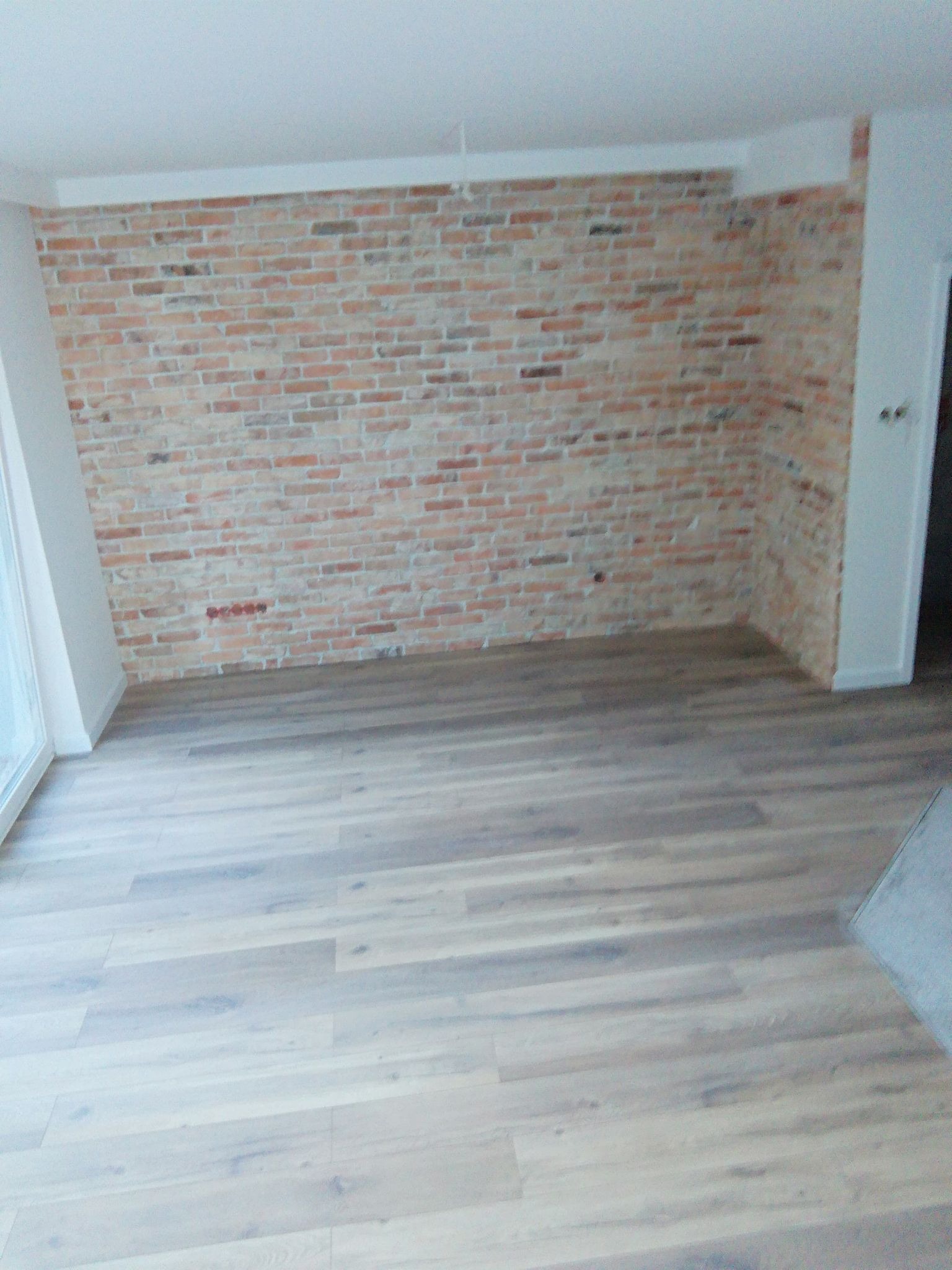 Wnętrze pokoju z wyeksponowaną ścianą z cegły w odcieniach czerwieni i beżu oraz jasnymi panelami podłogowymi z widoczną strukturą drewna.