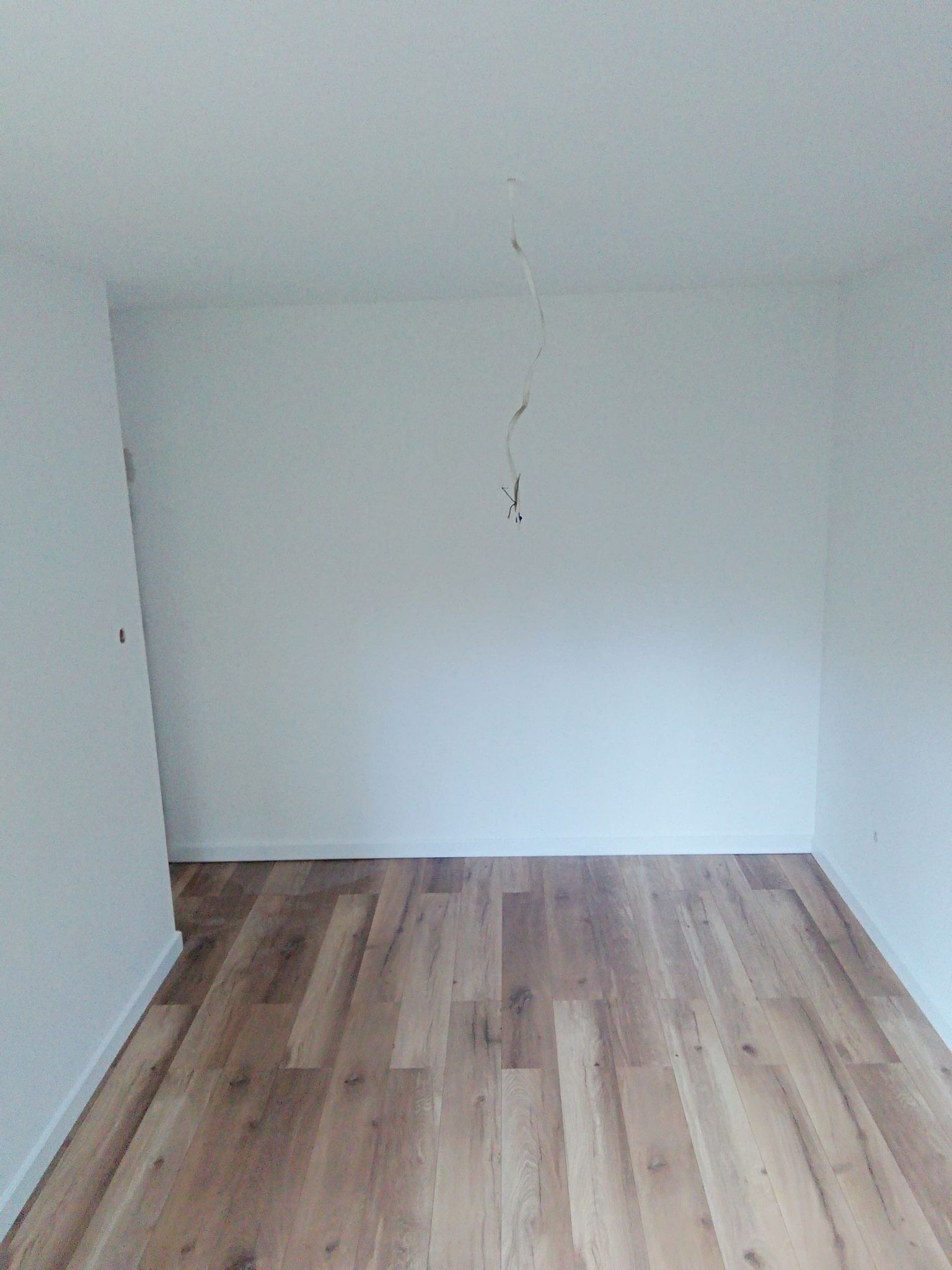 Pusty pokój po remoncie z drewnianą podłogą, świeżo pomalowanymi białymi ścianami i wystającym kablem z sufitu.