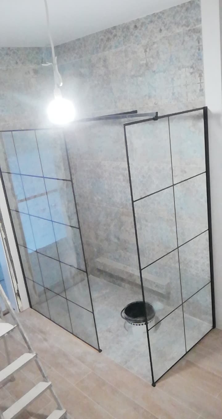 Nowoczesna kabina prysznicowa z czarnymi profilami i szklanymi panelami w kwadraty, zamontowana w łazience z płytkami imitującymi marmur, widoczny czarny kubeł na podłodze i fragment drabiny...