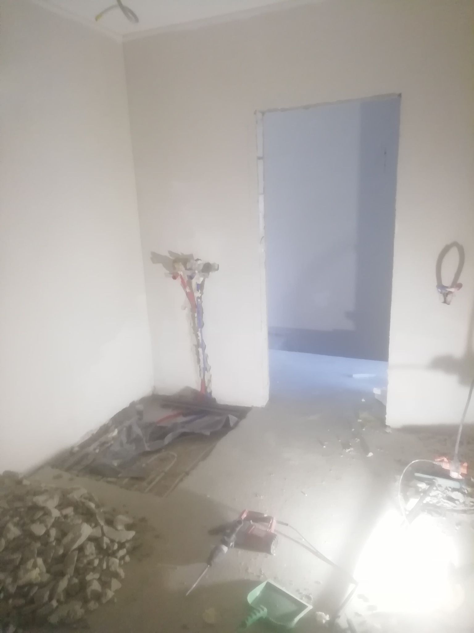 Remont łazienki w trakcie: odkryte rury wodne na ścianie, gruz na podłodze, widoczny młot udarowy i szpachelka, surowy stan pomieszczenia.