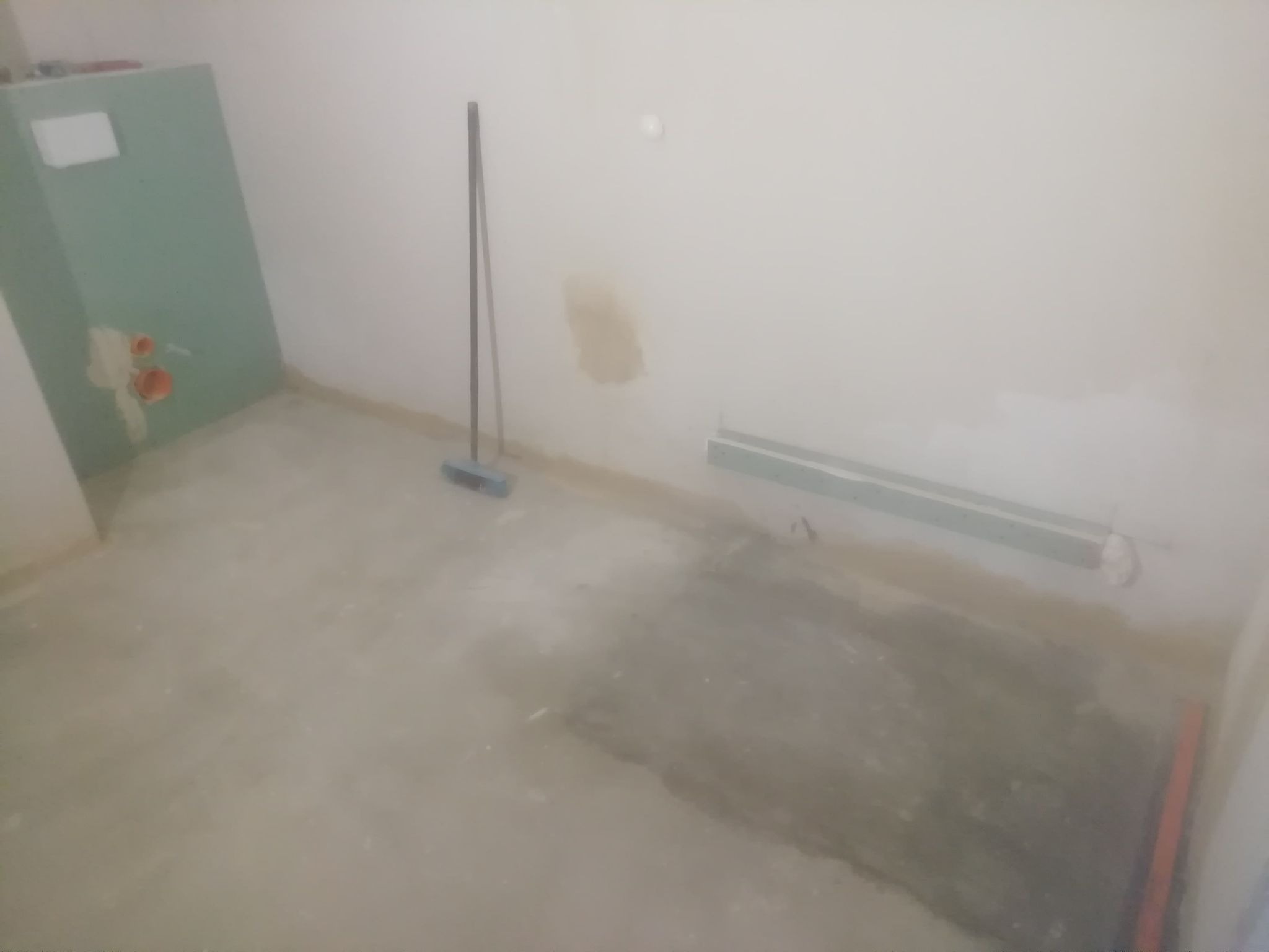 Surowy stan łazienki w trakcie remontu: widoczne ślady po gładzi na ścianach, niezabezpieczone rury, pionowa listwa przypodłogowa i płyta gipsowo-kartonowa z instalacją.