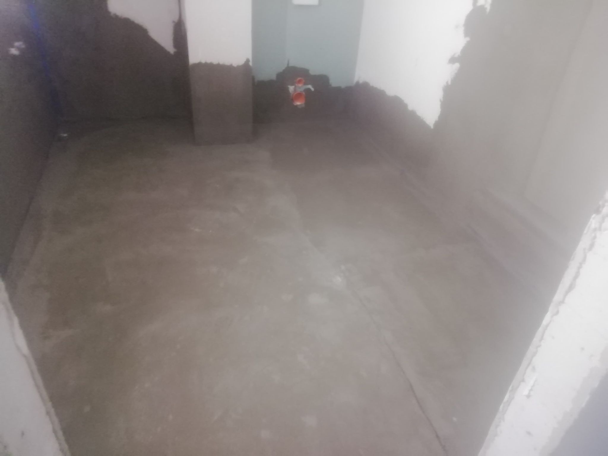 Przygotowanie podłoża w łazience: wyrównana wylewka podłogowa, ściany częściowo pokryte szarą zaprawą, widoczne rury instalacji wodnej.