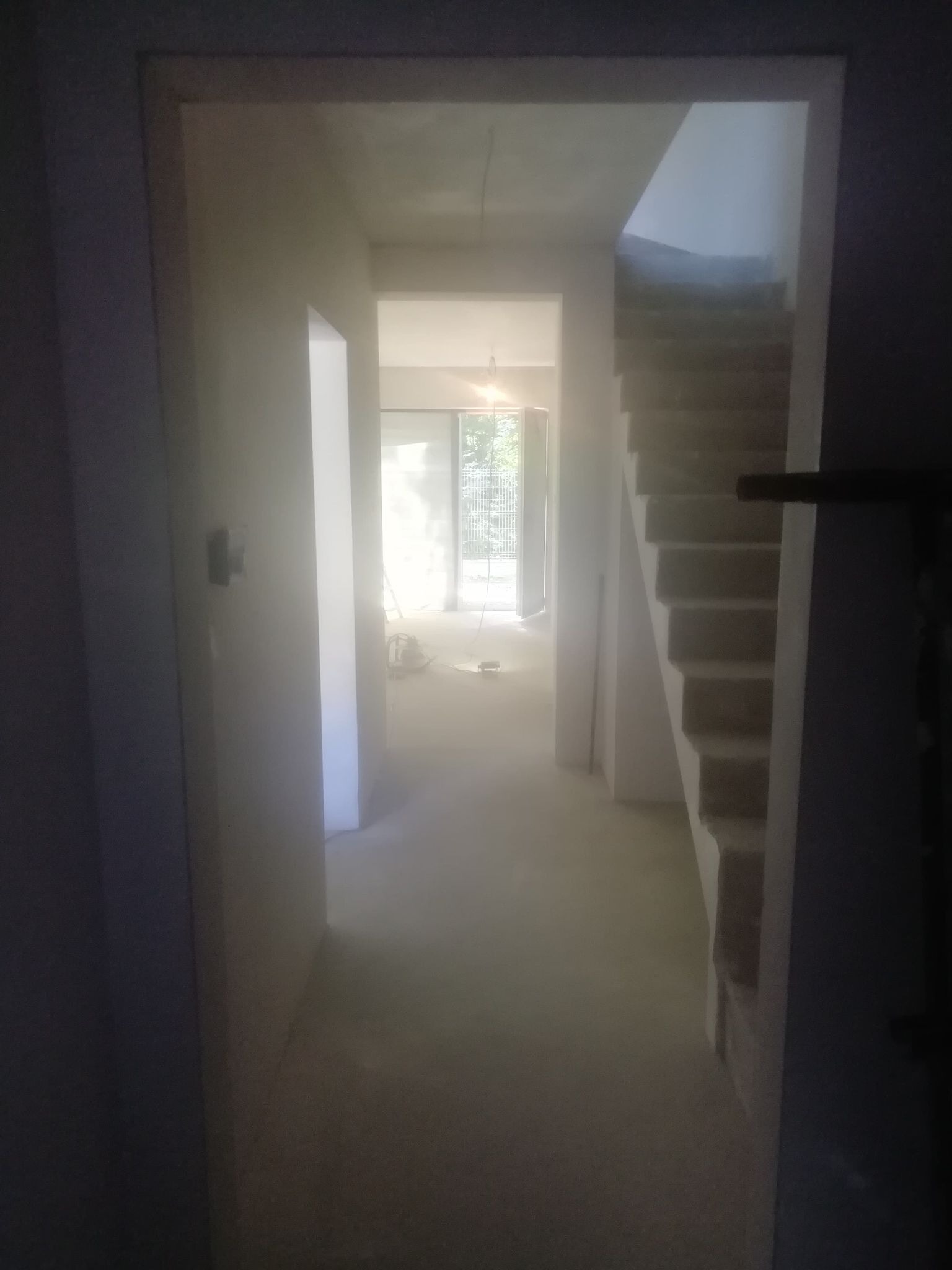 Wnętrze w trakcie remontu: widok na korytarz z surowymi ścianami, schodami i oknem na końcu, widoczne ślady prac budowlanych.