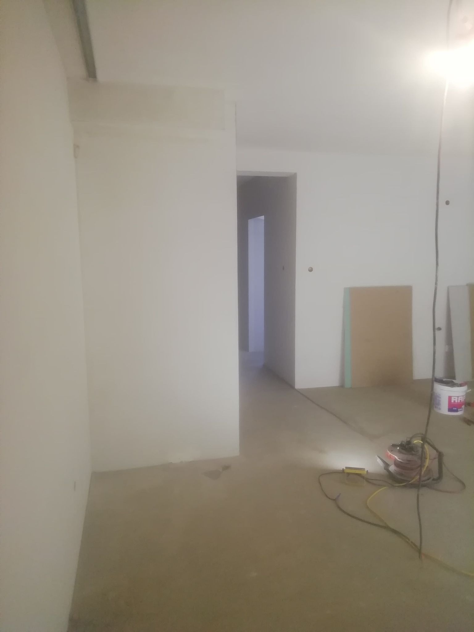 Wnętrze w trakcie prac wykończeniowych: gładkie, białe ściany, surowa podłoga, widoczne narzędzia i materiały budowlane, przejście do kolejnego pomieszczenia w tle.