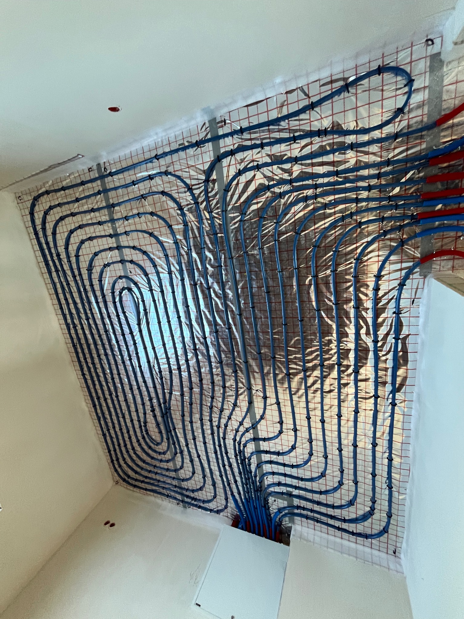 Niebieskie rury instalacji ogrzewania podłogowego rozłożone na srebrnej folii izolacyjnej z siatką, widok z dołu, fragment ściany i sufitu w tle.