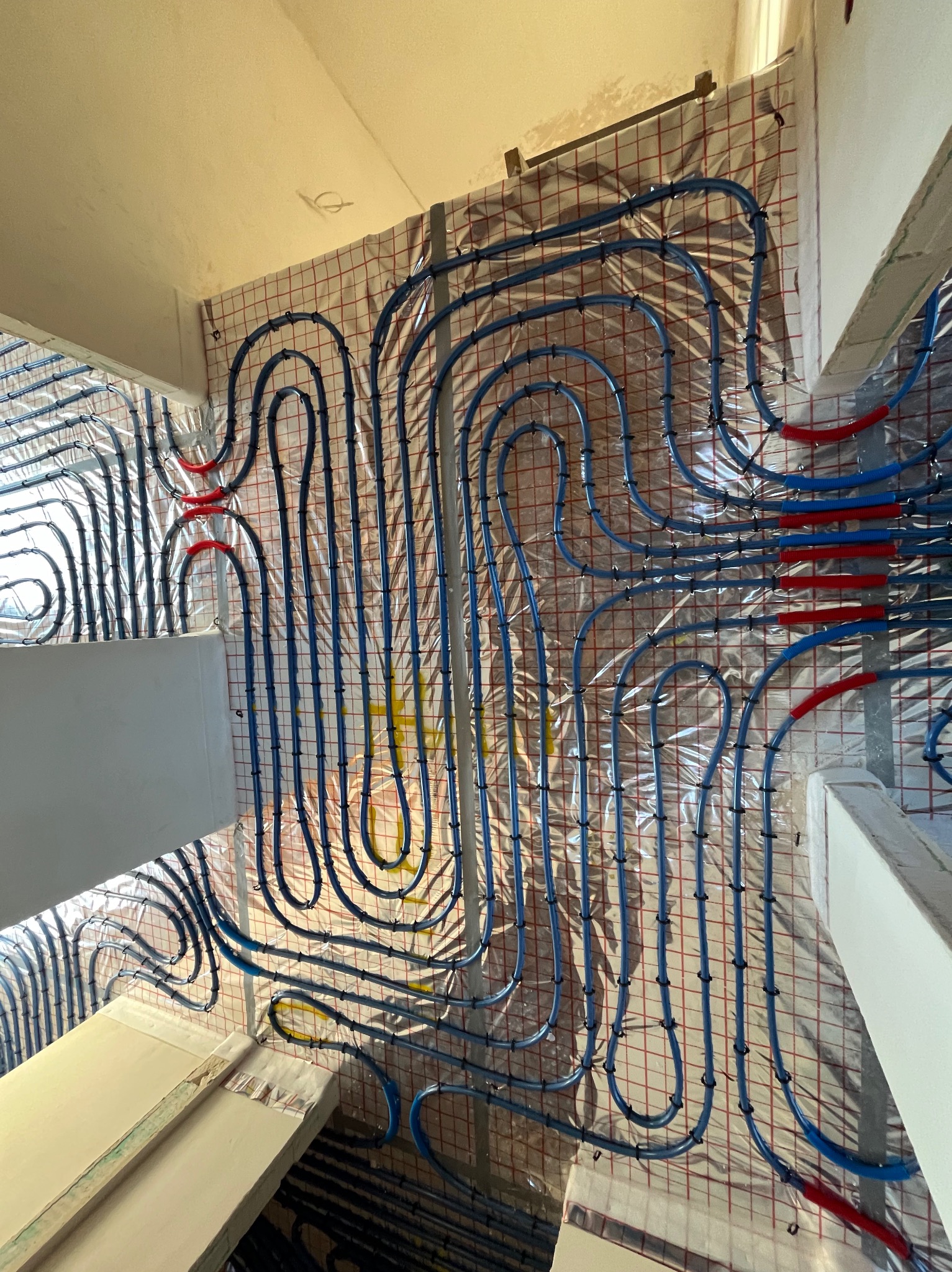 Montaż niebieskich rur systemu grzewczego na folii izolacyjnej z siatką, widoczne fragmenty ścian i sufitu w surowym stanie.