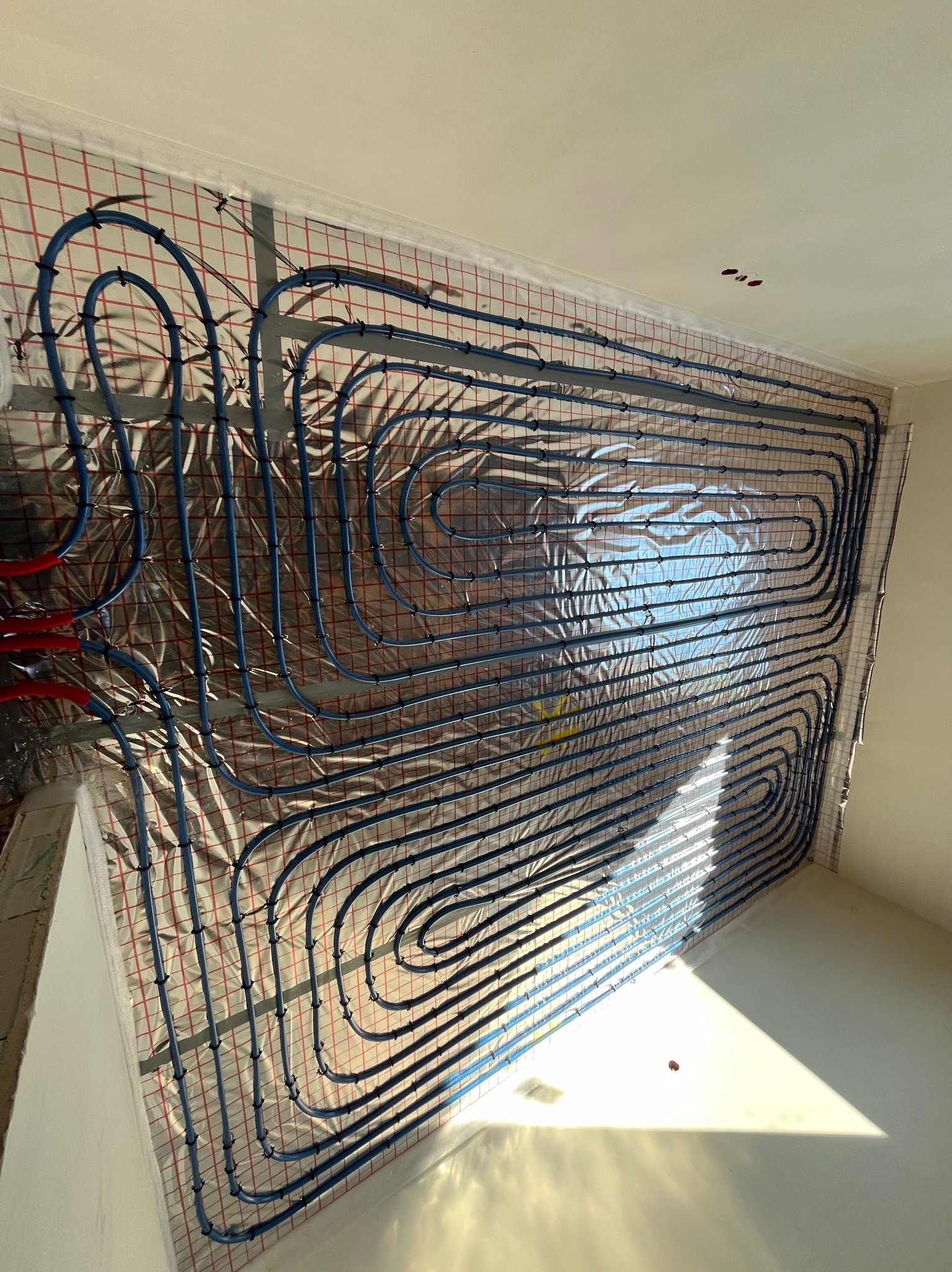 Niebieskie rurki instalacji grzewczej rozprowadzone na aluminiowej folii z czerwoną siatką, zamocowane czarnymi klipsami, widok z góry na surową wylewkę podłogową w słonecznym pokoju.