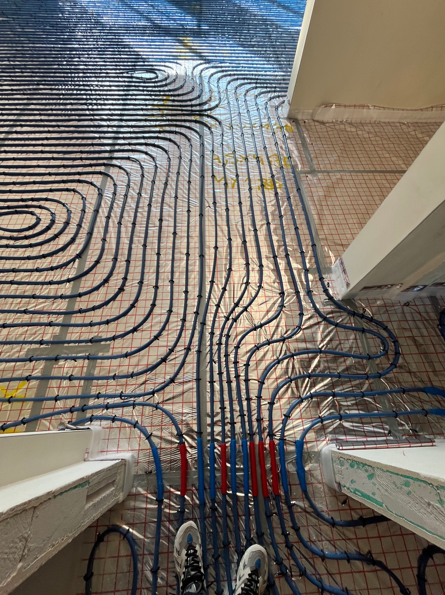 Montaż ogrzewania podłogowego: niebieskie rurki ułożone na folii z czerwoną siatką, widoczne fragmenty ścian i para butów roboczych w dolnej części kadru.