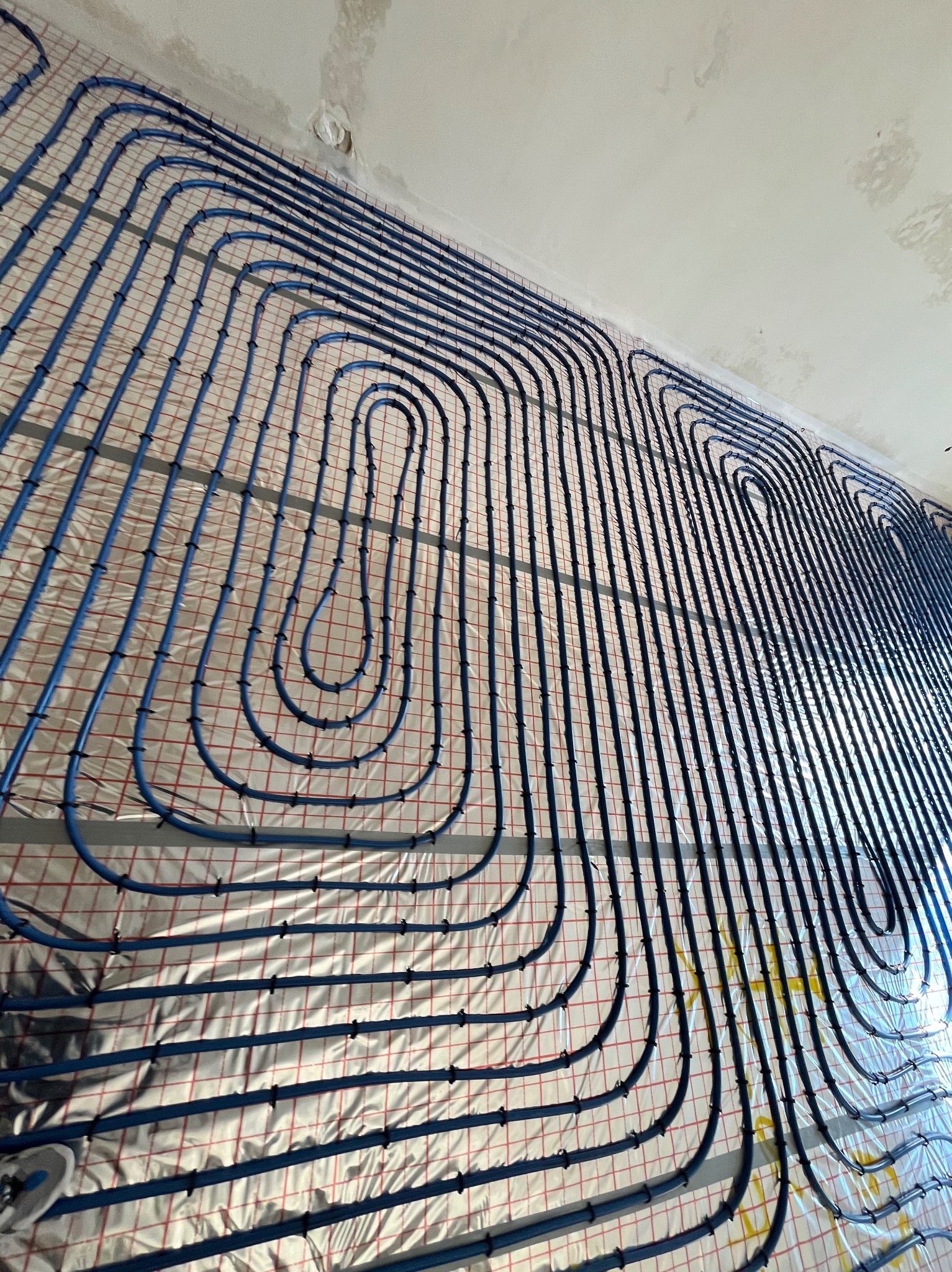 Niebieskie rury układane w pętle na folii z nadrukowaną siatką, przygotowanie do instalacji podłogowej, widok z dołu na sufit z widocznymi niedoskonałościami.