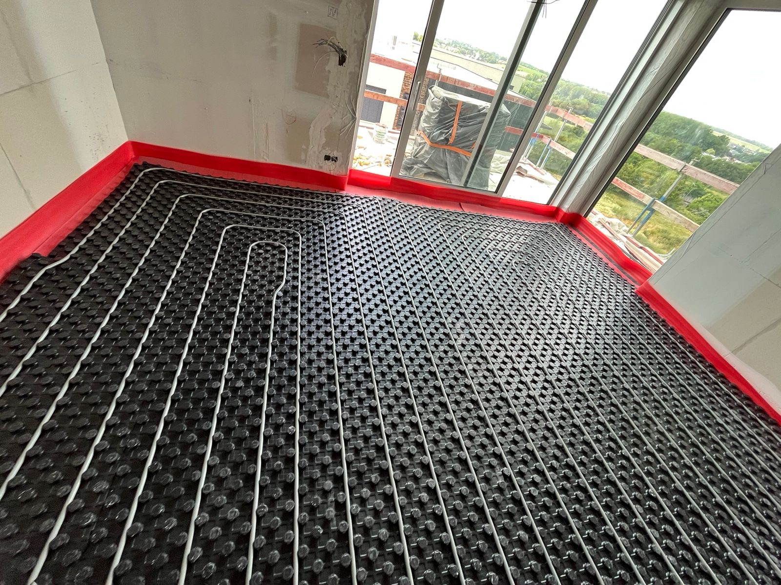Rozłożona instalacja ogrzewania podłogowego z czarną matą i białymi rurkami w surowym pomieszczeniu z dużym oknem i widokiem na zieleń.
