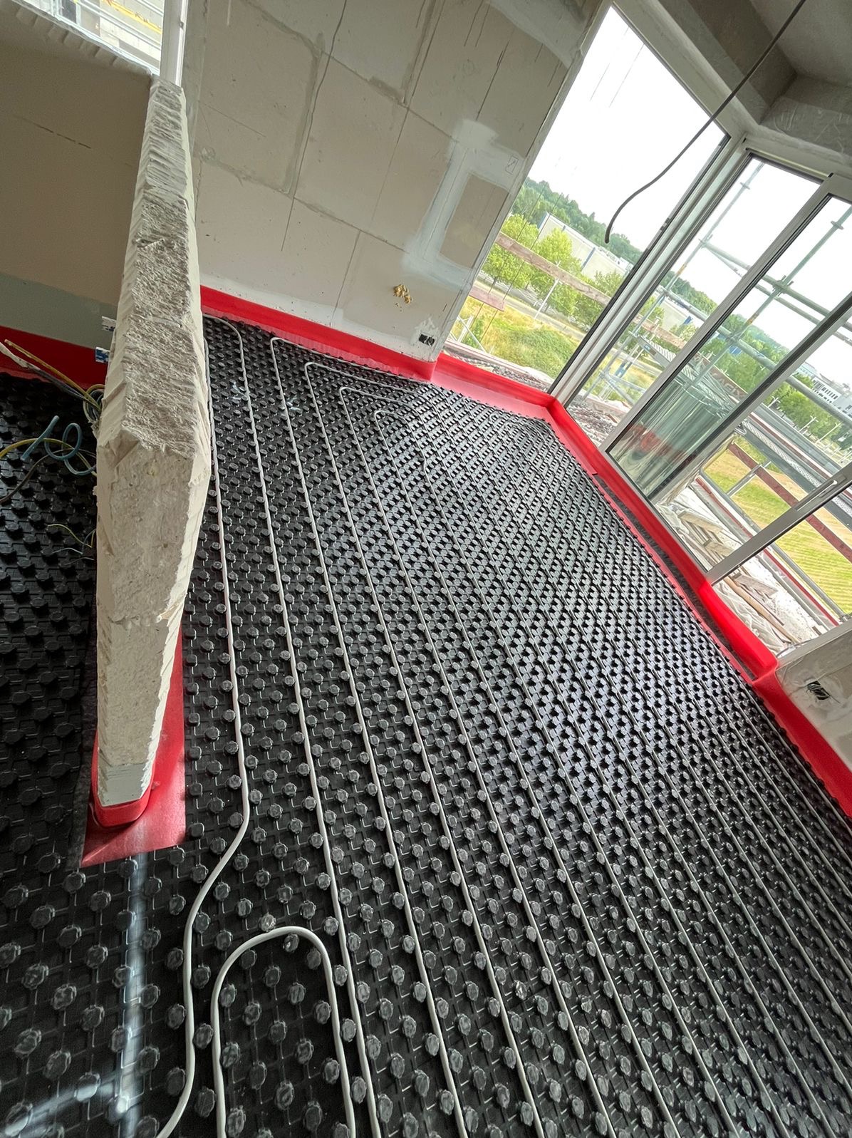 Montaż instalacji ogrzewania podłogowego w surowym pomieszczeniu, widoczne rurki układane na czarnej macie z wypustkami, czerwona taśma brzegowa, surowe ściany i widok na zewnątrz przez duże okno.